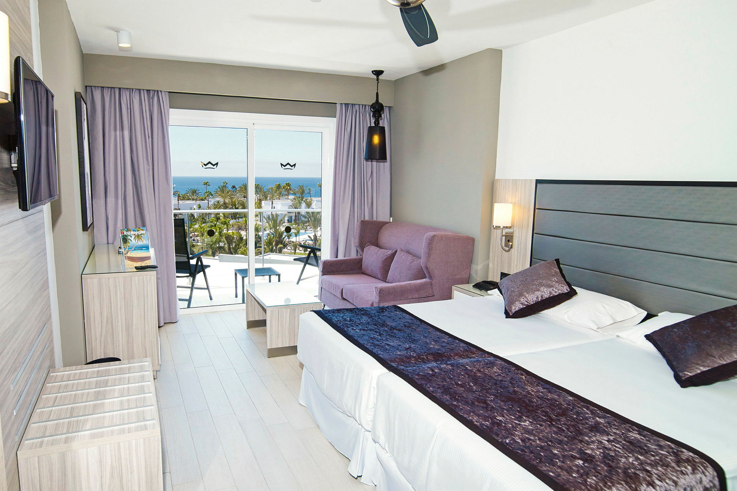 Modernt hotellrum med en dubbelsäng, soffgrupp och balkong med utsikt över havet. (AI-genererad text)
