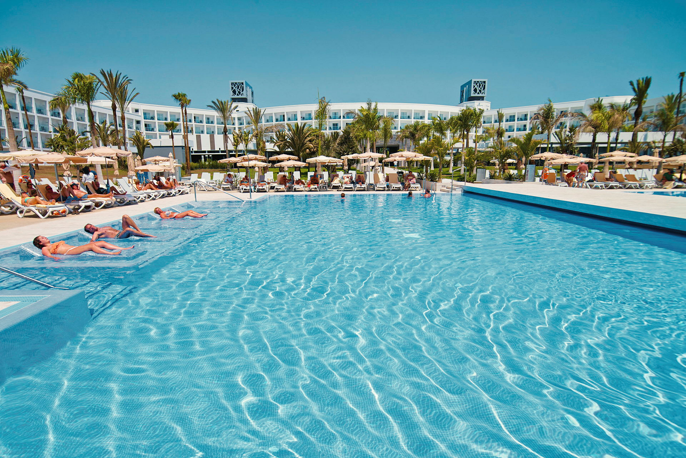 En stor pool omgiven av solstolar och parasoller, med ett hotell i bakgrunden. (AI-genererad text)
