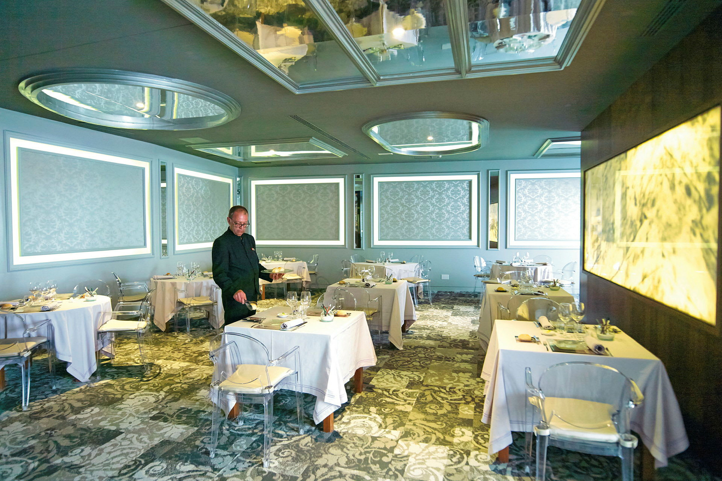 En elegant restaurang med bord klädda i vita dukar, mönstrade väggar och moderna ljusinstallationer. (AI-genererad text)