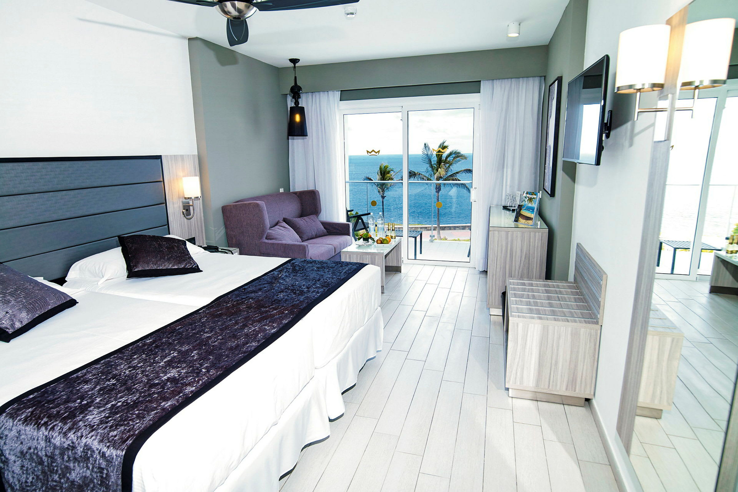 Hotellrum med dubbelsäng, soffa och havsutsikt utanför stora fönster. (AI-genererad text)