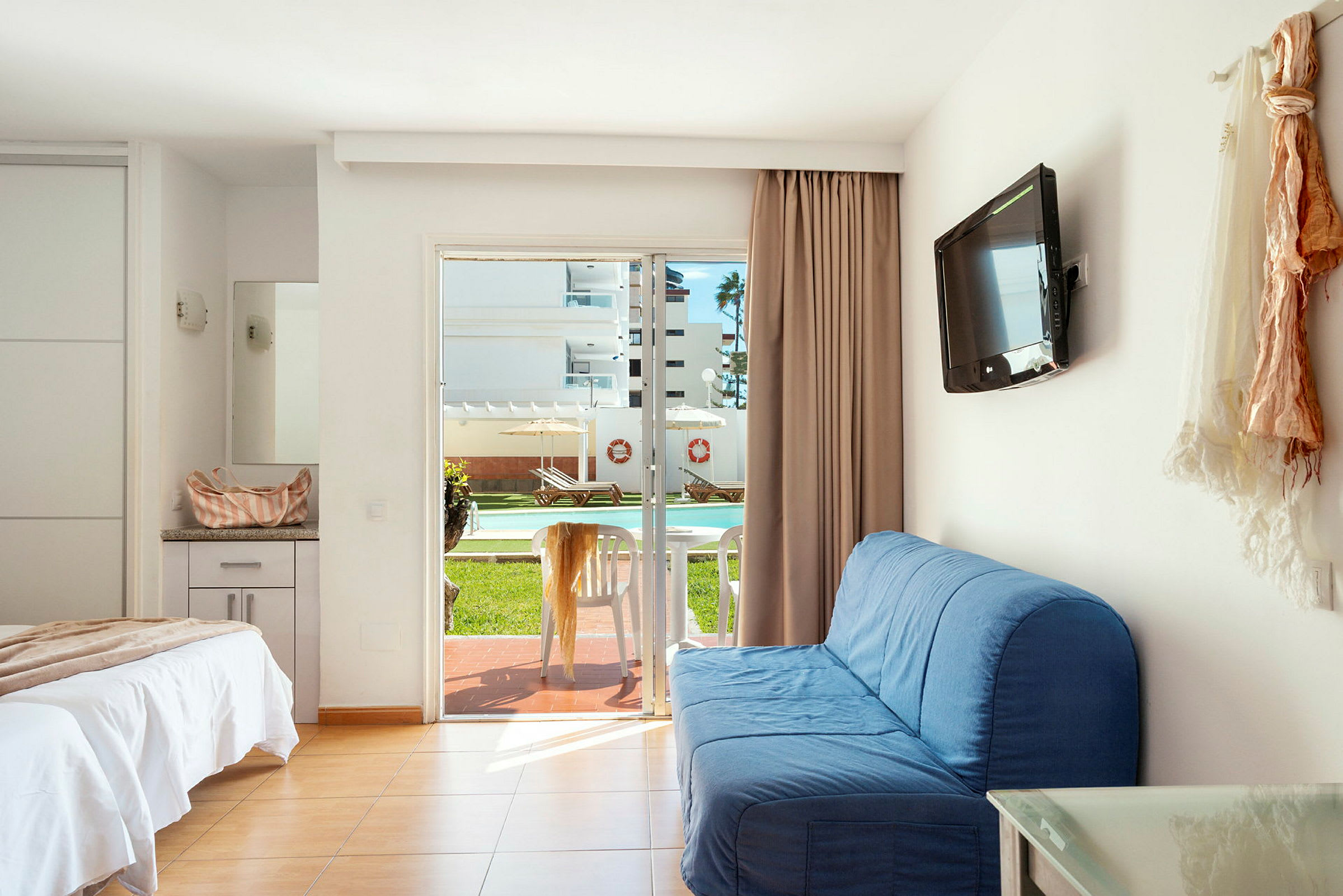 Ett hotellrum med en bäddad säng, blå soffa, väggmonterad TV och en glasdörr som leder ut till en terrass med bord och stolar. Utsikten visar en utomhuspool och solstolar. (AI-genererad text)