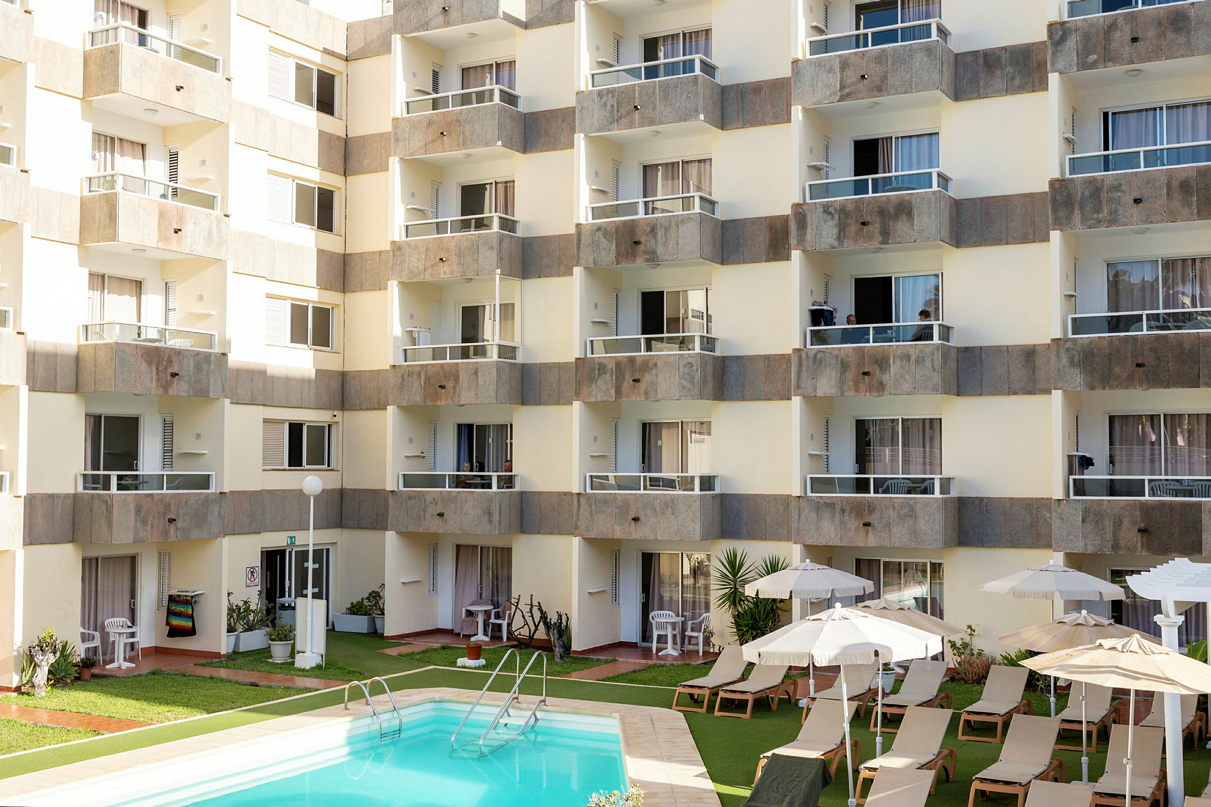 Bild av en hotellbyggnad med flera balkonger runt en gård som har en pool och solstolar med parasoller. (AI-genererad text)