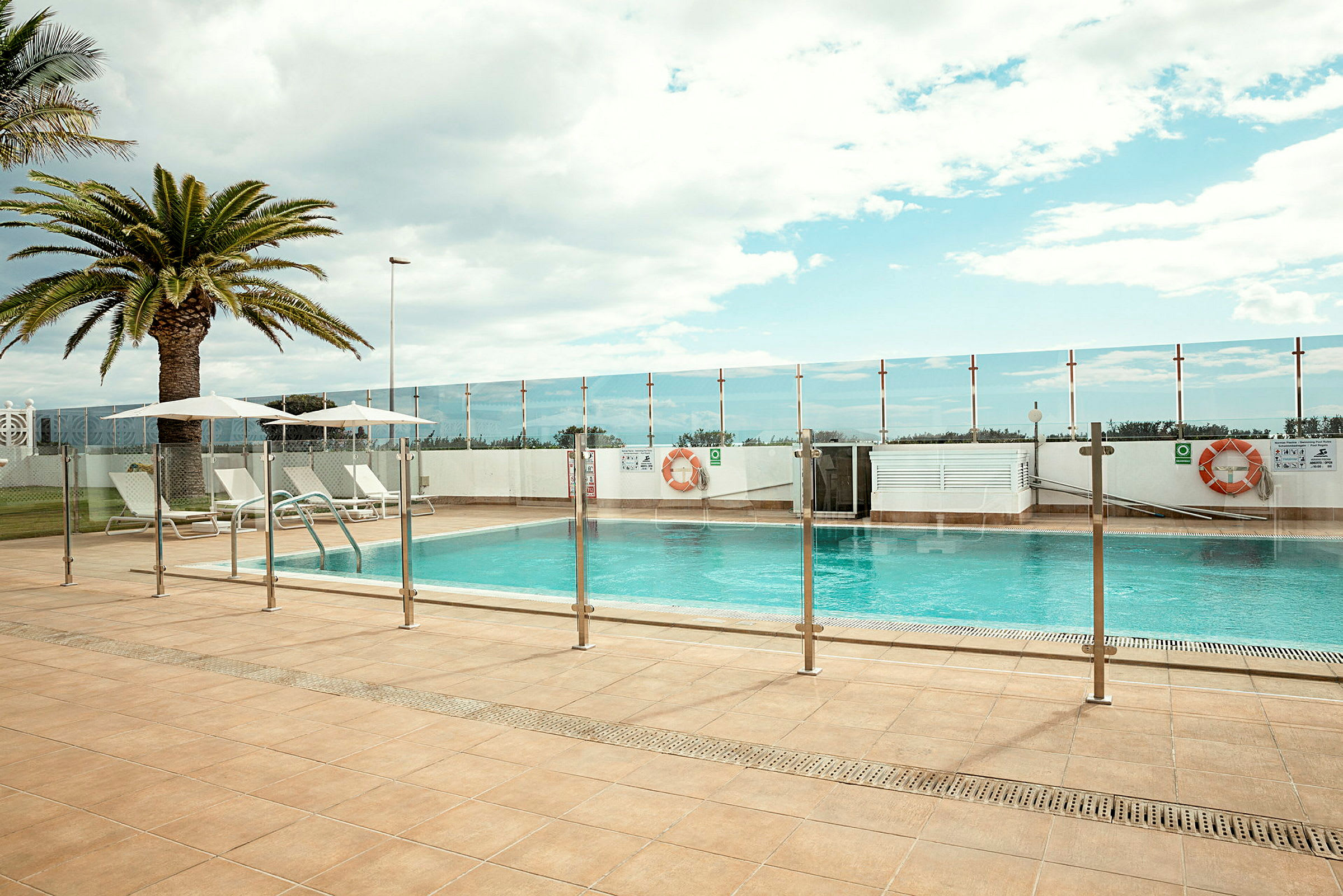 Utomhuspool med solstolar och parasoll bredvid. Palmträd och glasstaket runt poolområdet. (AI-genererad text)