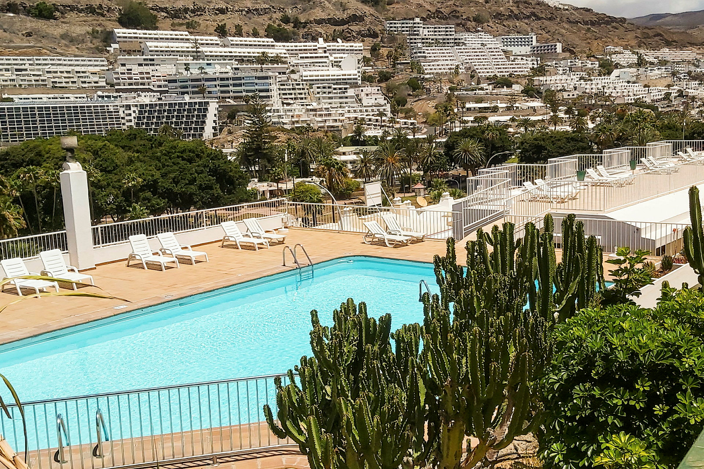 Utomhuspool med solstolar och grönska, omgiven av byggnader och berg. (AI-genererad text)