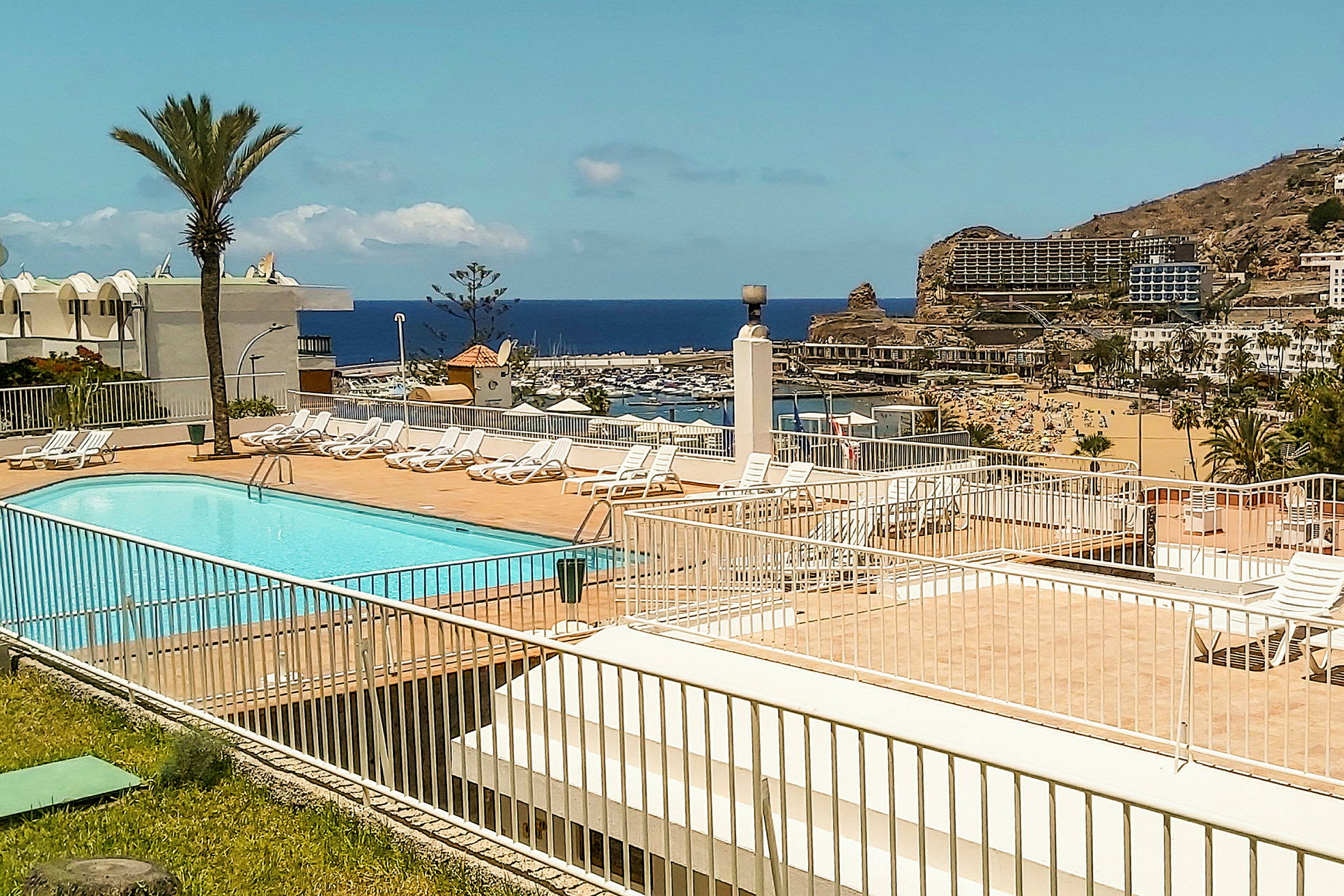 En pool med solstolar omgiven av ett staket, med en utsikt över en strand och havet i bakgrunden. (AI-genererad text)