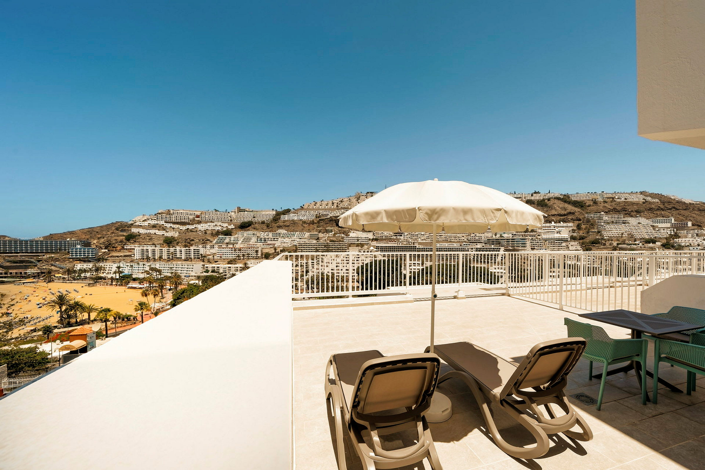 Terrass med solstolar och parasoll, utsikt över en strand och stad mot berg i fjärran. (AI-genererad text)