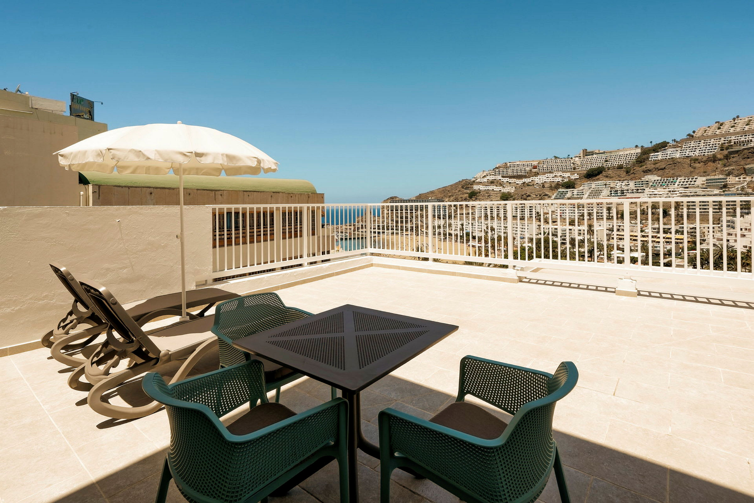 Utsikt från en terrass med bord, stolar och solstolar, mot havet och byggnader på bergssluttning. (AI-genererad text)