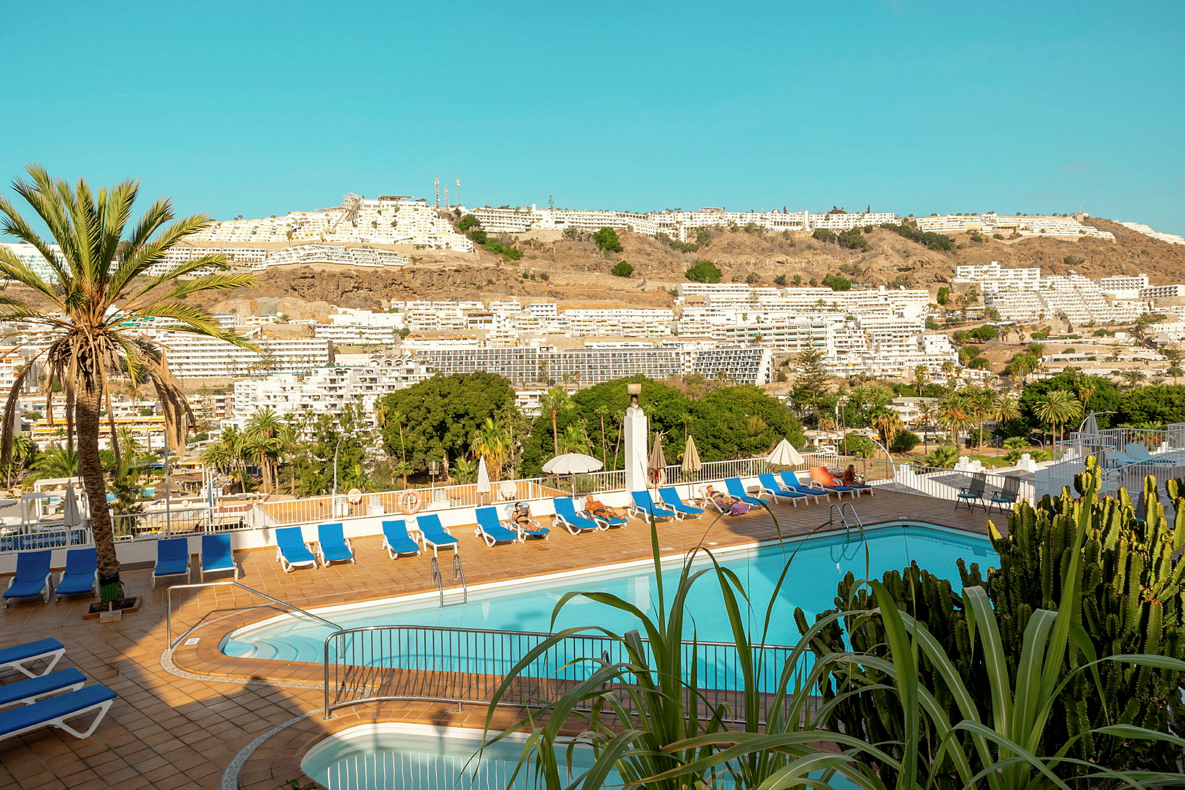 Bild på en hotellpool med solstolar och parasoller. I bakgrunden syns byggnader och grönska. (AI-genererad text)