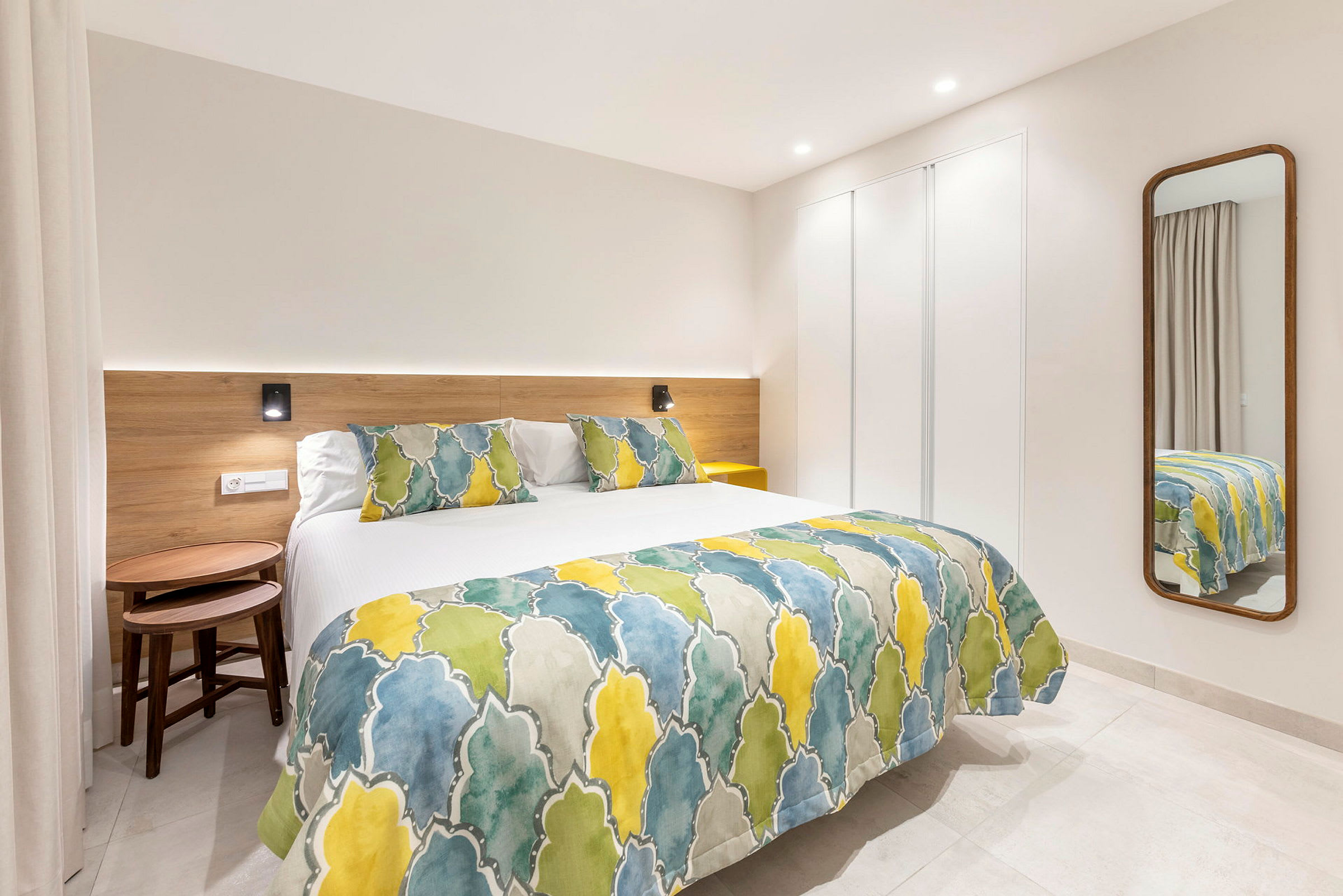 Ett modernt hotellrum med en säng klädd i färgglatt överkast och kuddar samt en spegel på väggen. (AI-genererad text)