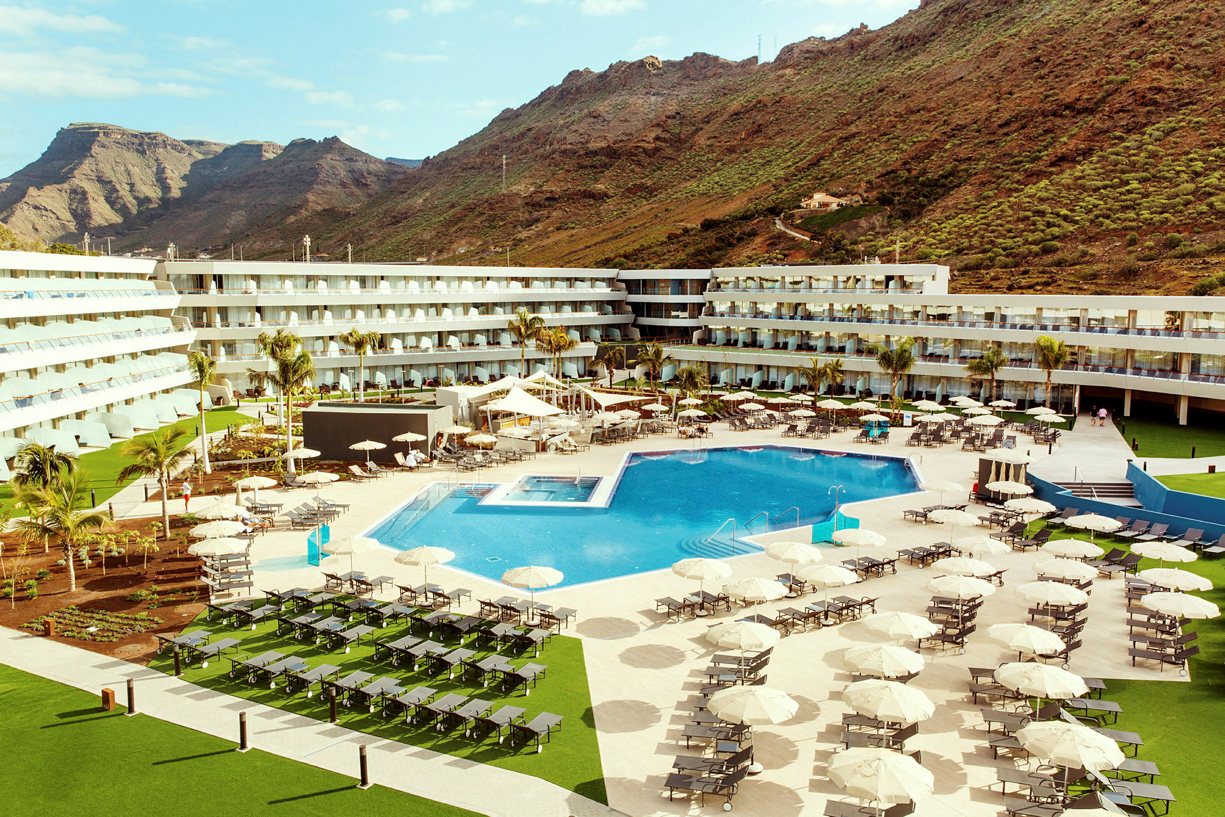 Ett hotellområde med en stor utomhuspool, solstolar och parasoller, omgivet av byggnader och berg i bakgrunden. (AI-genererad text)