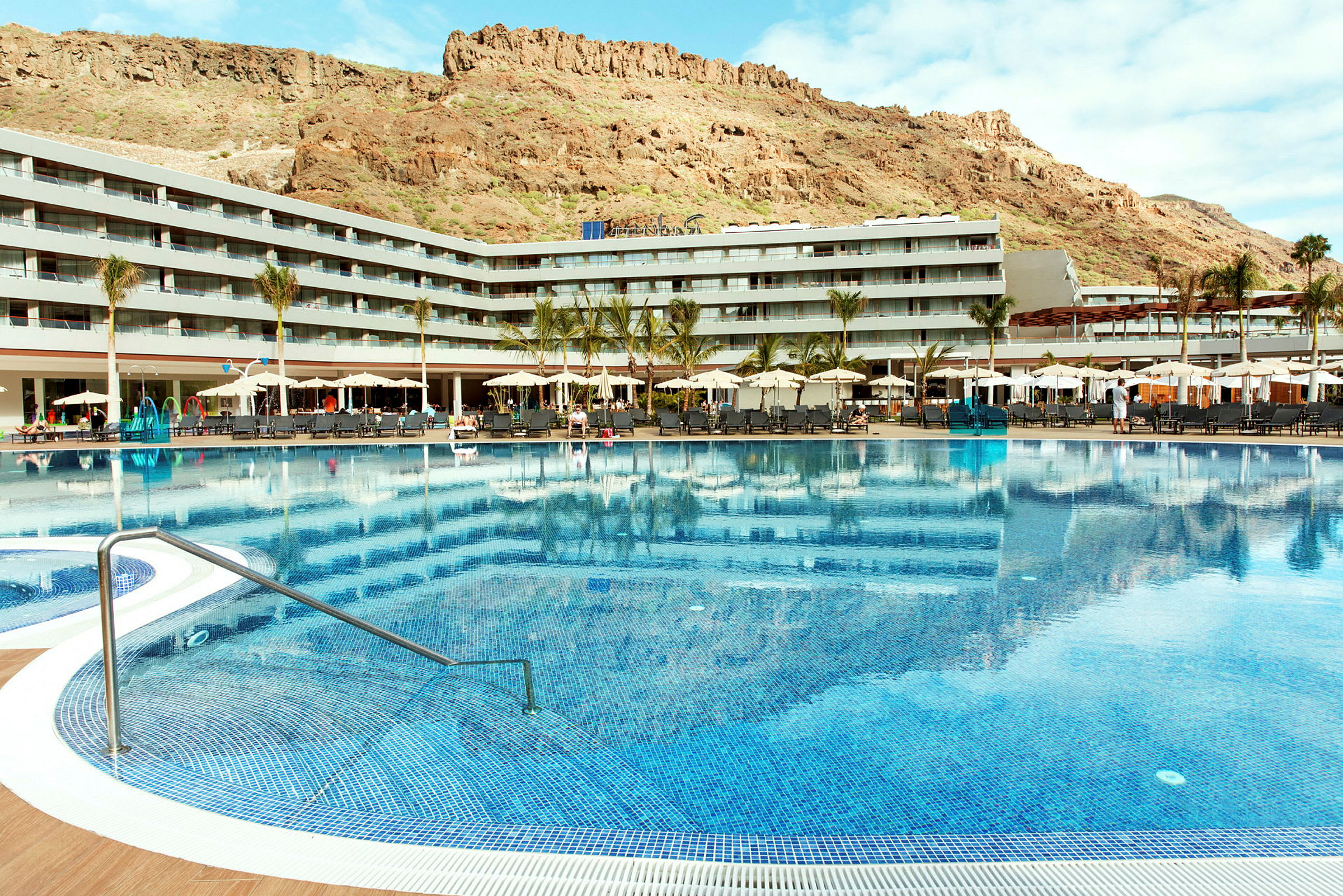 Stor utomhuspool framför hotellbyggnad och berg i bakgrunden. (AI-genererad text)