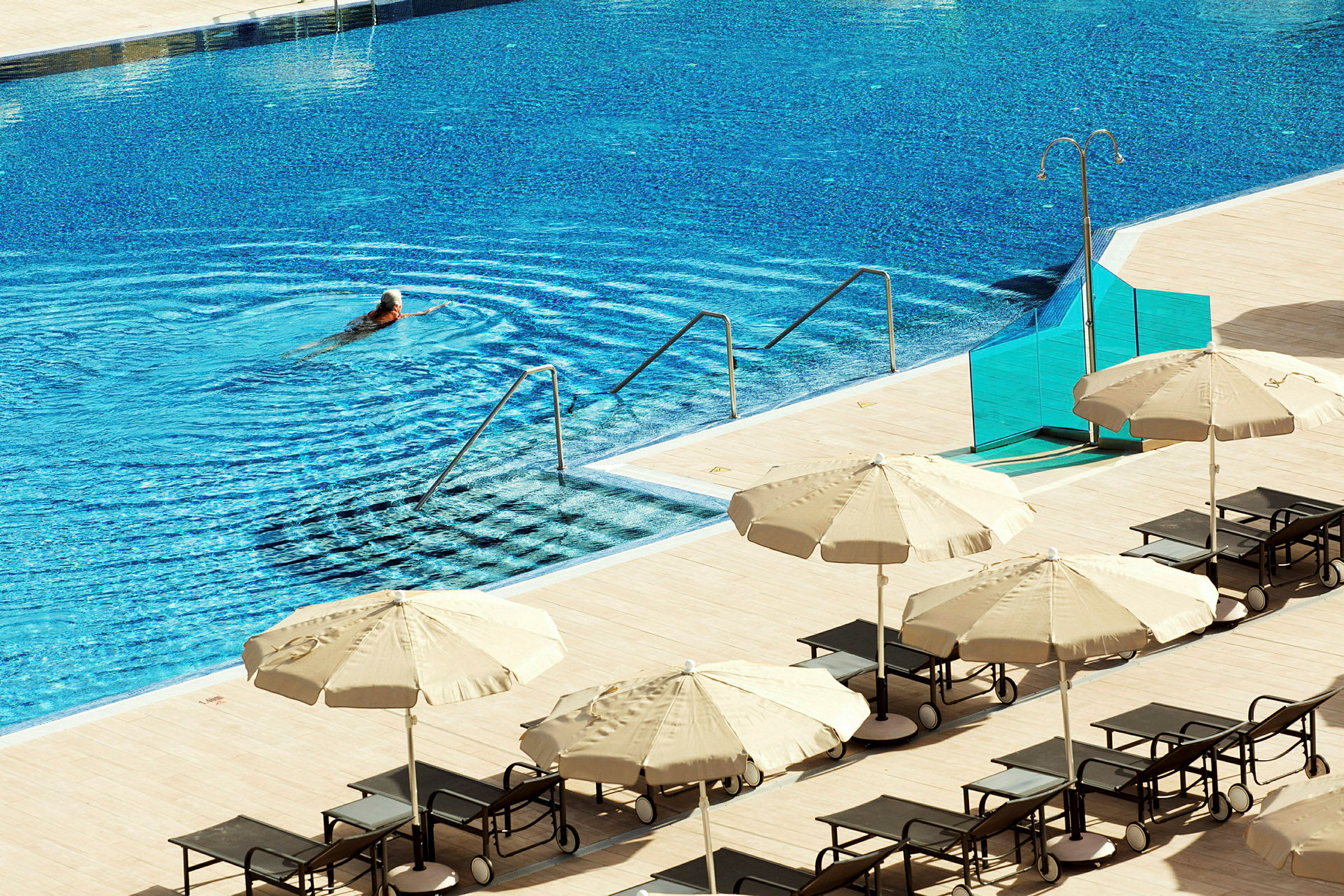 En person simmar i en swimmingpool med solstolar och parasoll vid sidan. (AI-genererad text)