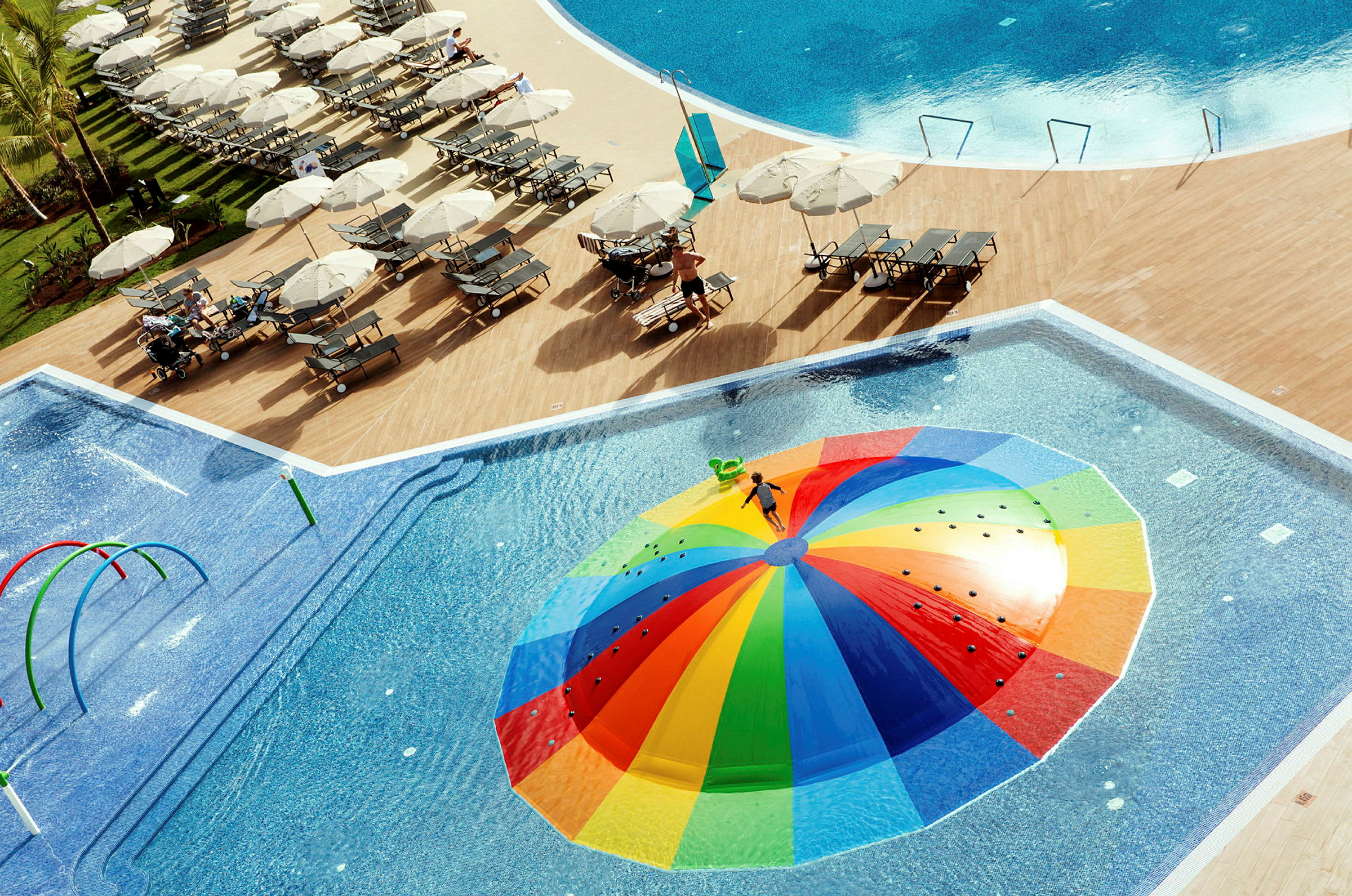 Utsikt över en pool med ett färgglatt vattenlekredskap vid en resort, omgiven av solstolar och parasoller. (AI-genererad text)