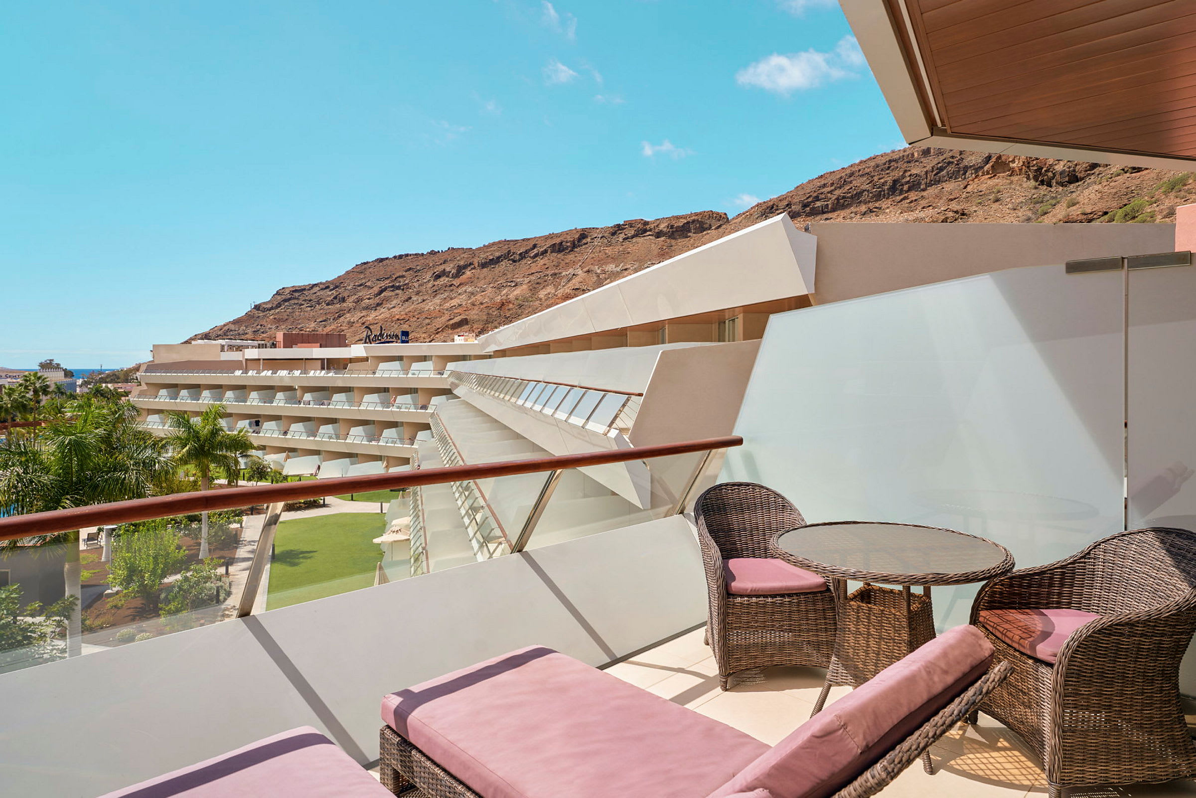 En terrass med solstolar och möbler, med utsikt över hotellbyggnader, palmträd och berg. (AI-genererad text)