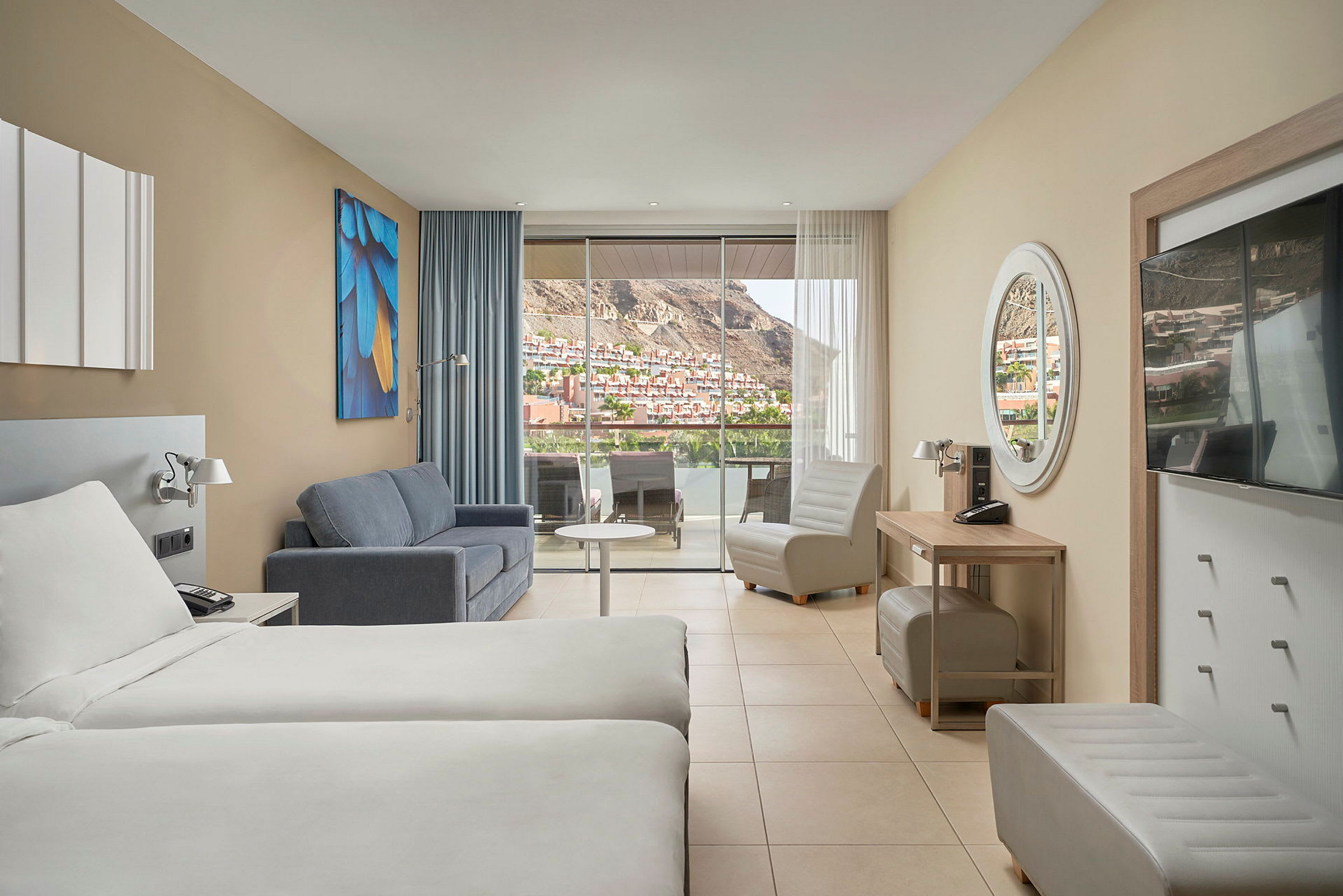 Hotellrum med två sängar, soffa, fåtölj och stor balkong med utsikt över en bergig stadsdel. (AI-genererad text)