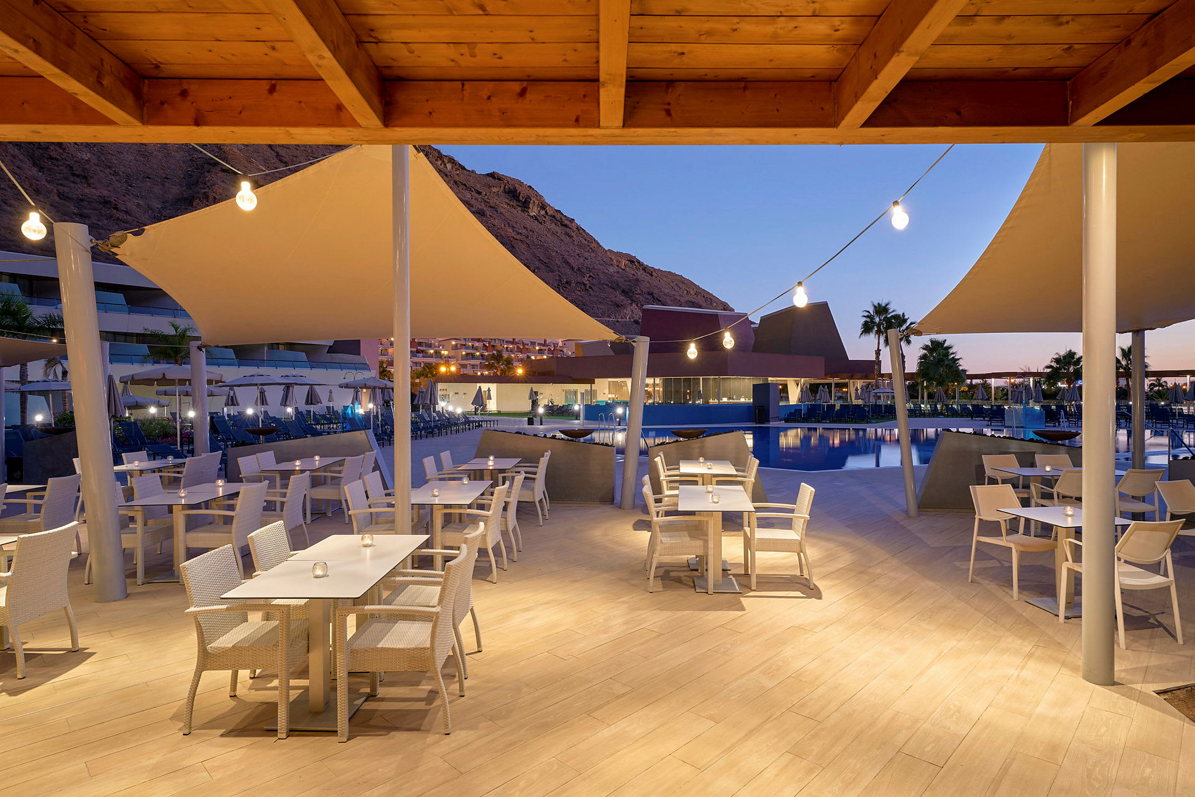 Utomhusrestaurang med vita stolar och bord vid en pool, kvällsbelysning och berg i bakgrunden. (AI-genererad text)