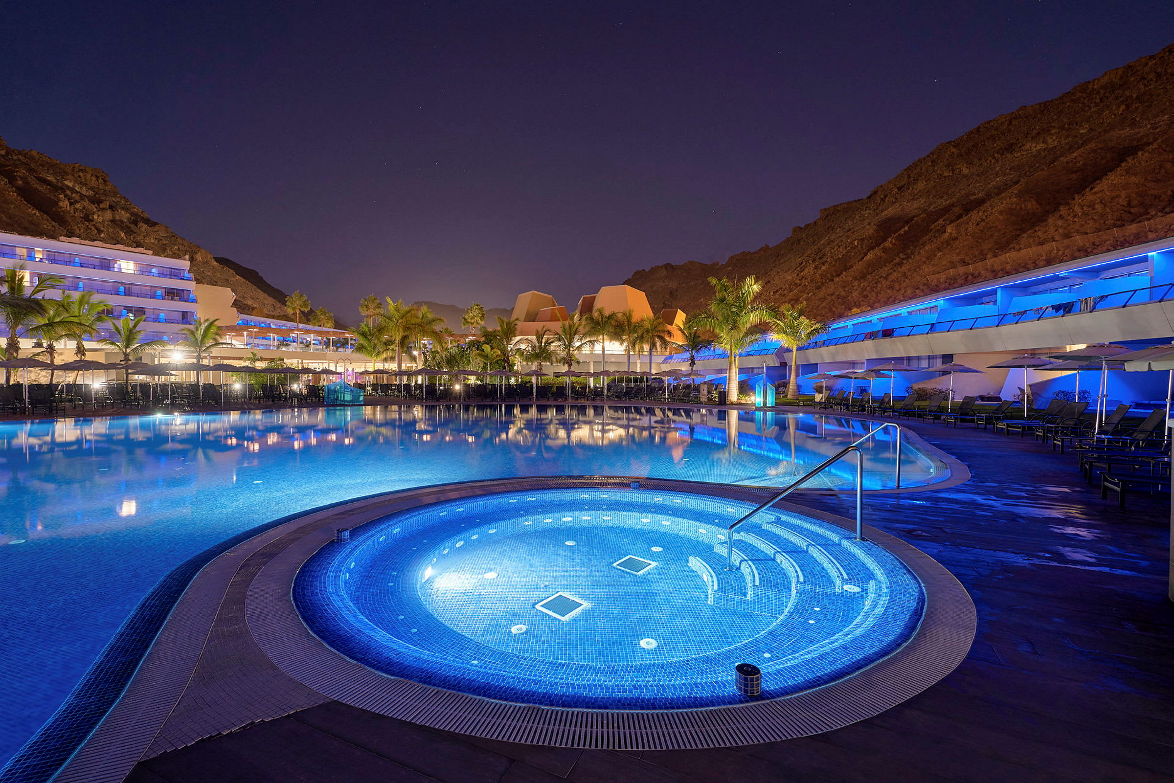 Bild av en upplyst poolområde med en mindre bubbelpool och omgivande hotellkomplex i bakgrunden under natthimmel. (AI-genererad text)