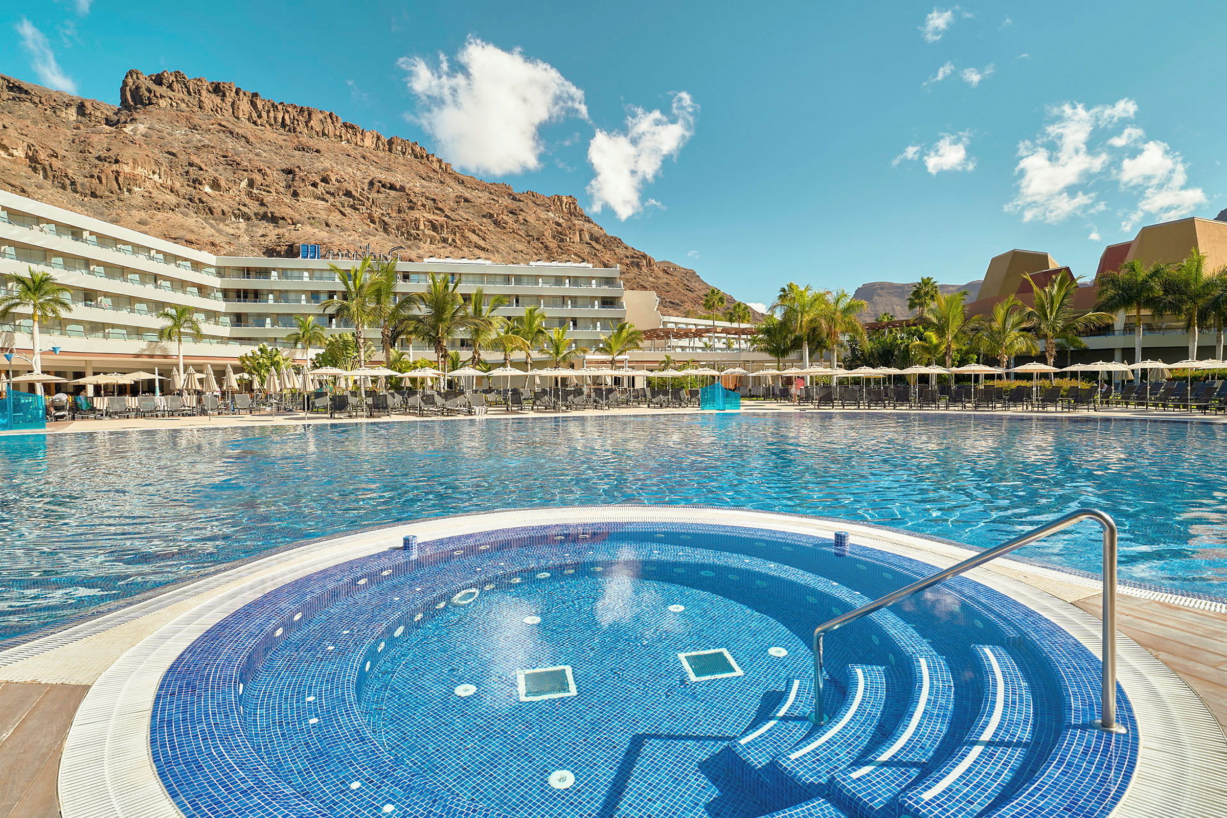 Hotellets poolområde med en mindre rund pool i förgrunden och omgivande byggnader samt berg i bakgrunden. (AI-genererad text)