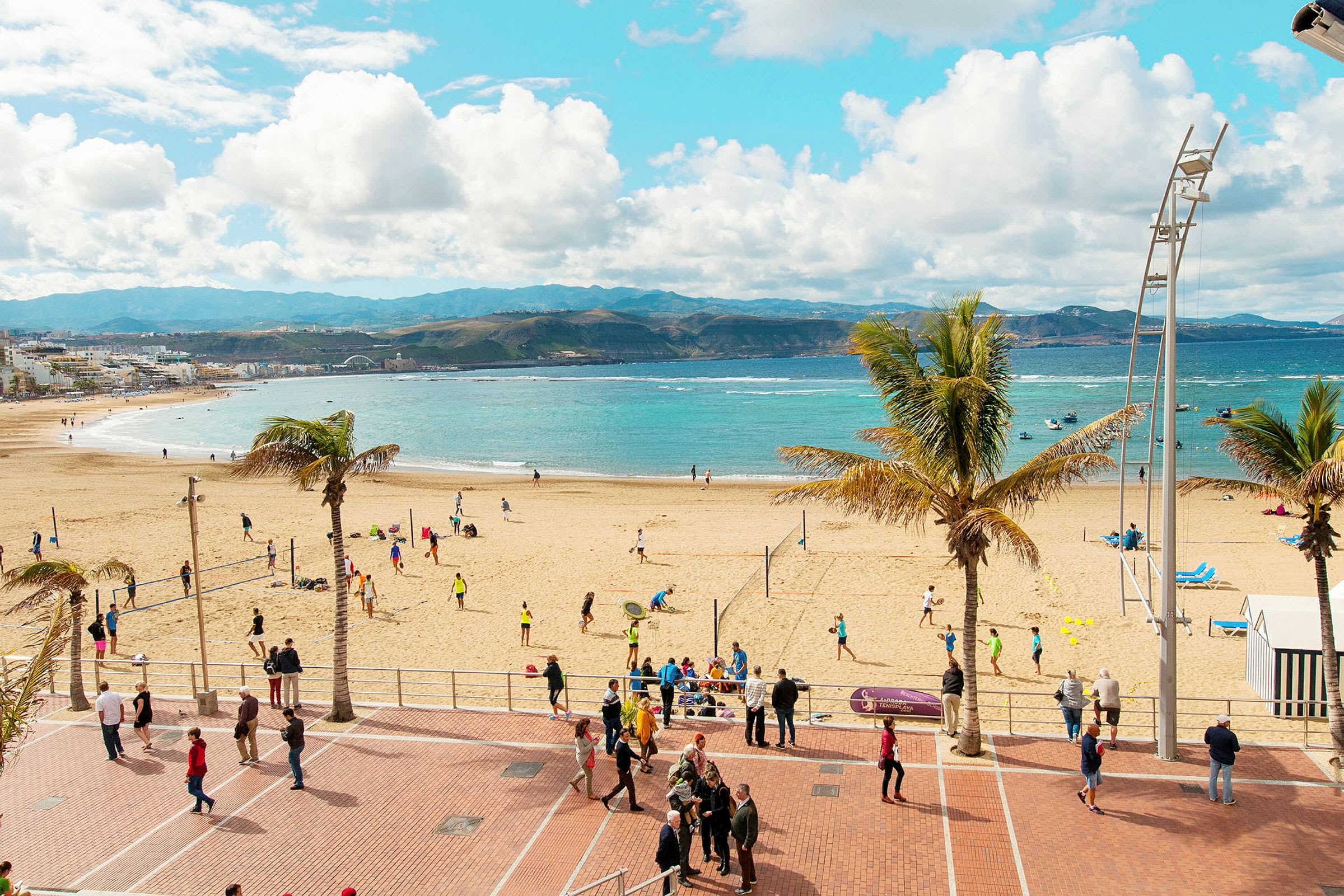 En strand med människor som spelar volleyboll och promenerar på strandpromenaden. Havet och bergen syns i bakgrunden. (AI-genererad text)