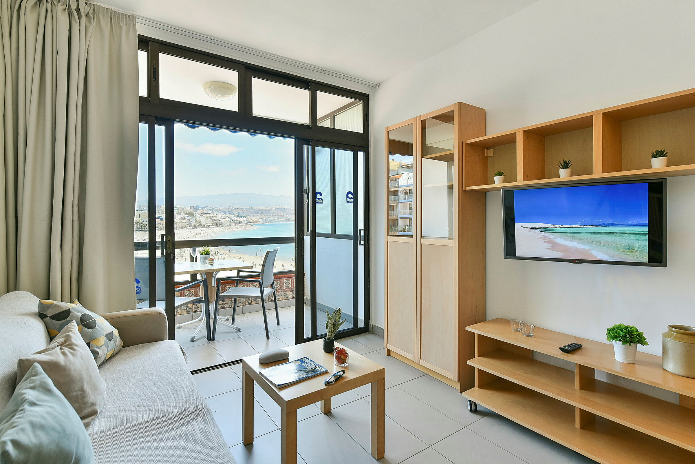 Ett vardagsrum med utgång till en balkong som har utsikt över en strand och havet. Rummet har möbler som en soffa, ett soffbord och en TV. (AI-genererad text)