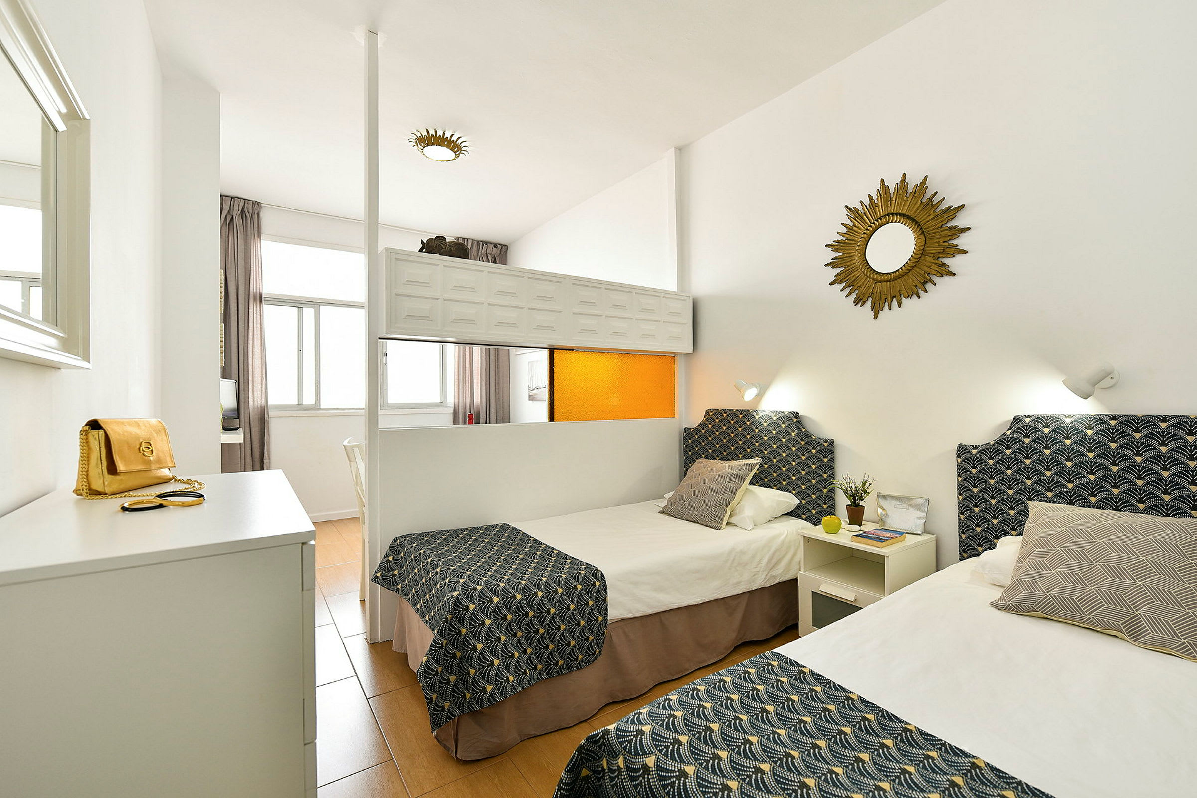 Ett hotellrum med två enkelsängar dekorerade med mönstrade överkast och kuddar, vita väggar och en spegel på väggen. (AI-genererad text)