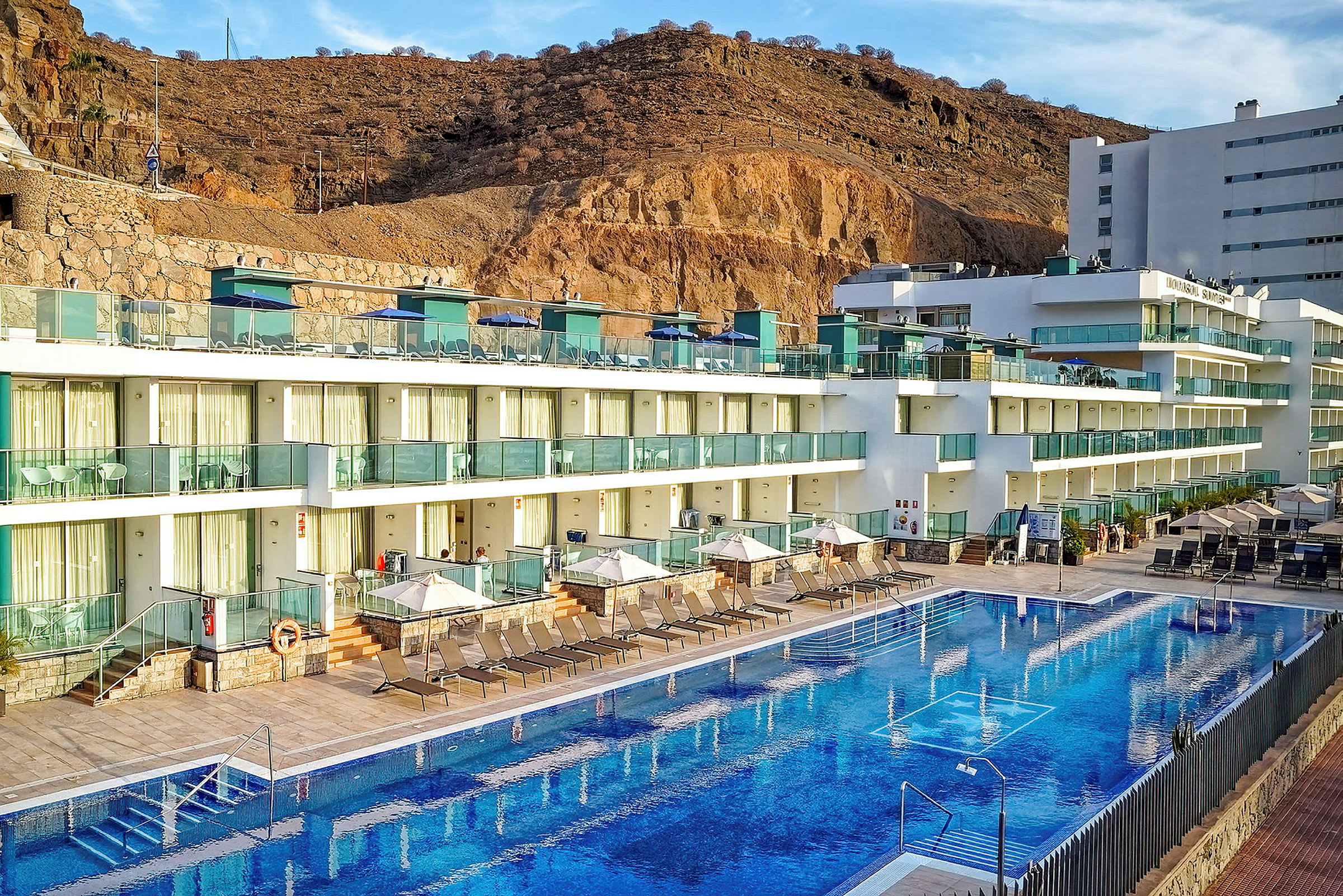 Hotellkomplex med flera våningar och glasfasader, solstolar samt parasoller vid en utomhuspool, omgiven av berg. (AI-genererad text)