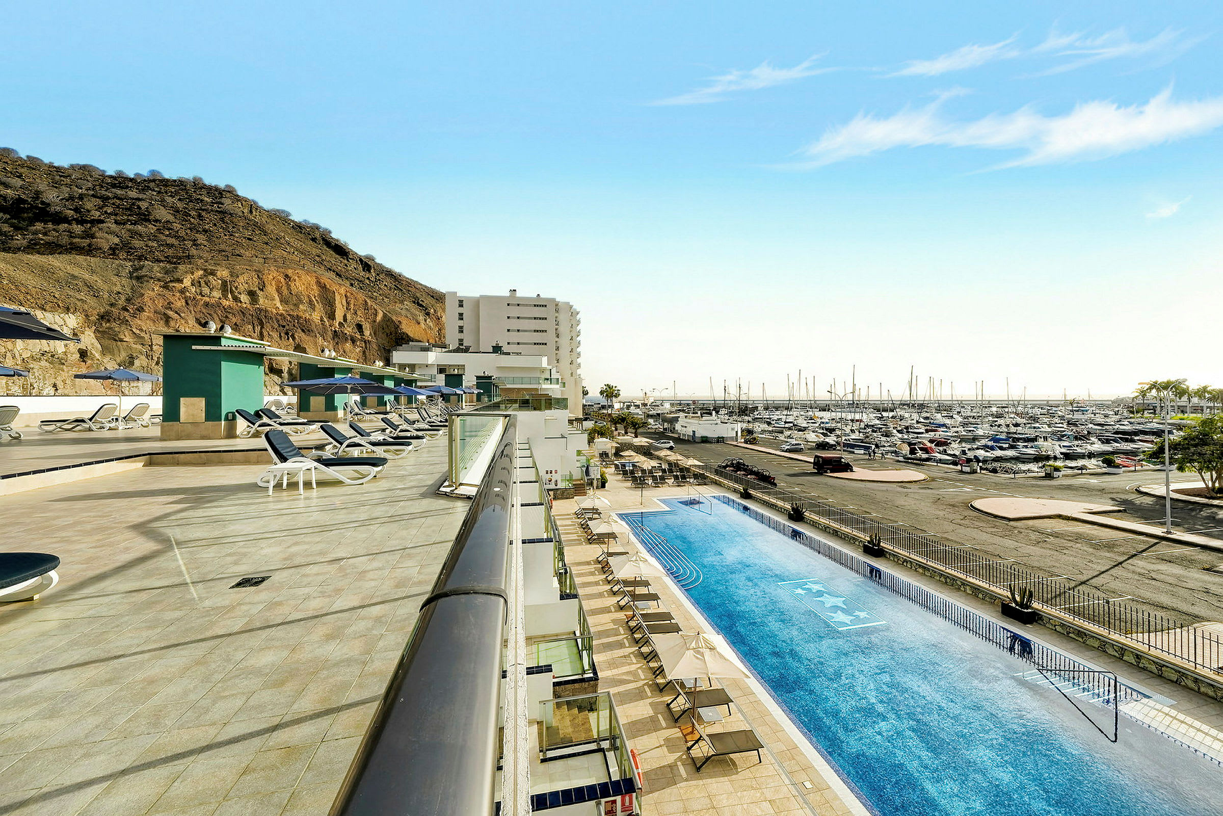 Poolområde med solsängar intill en marina och en bergssluttning. (AI-genererad text)