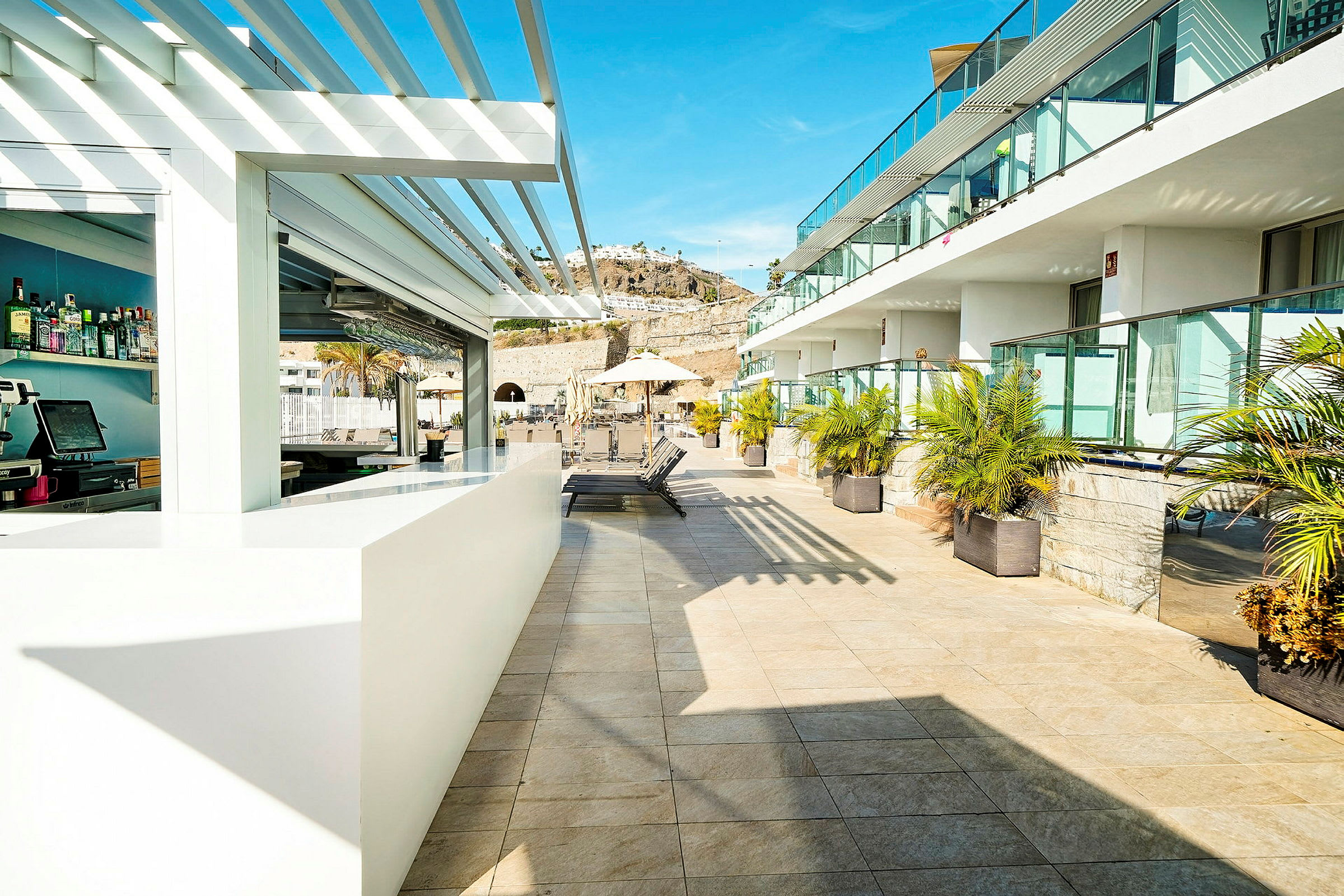 Bild av en solig hotellterrass med ett barområde och solstolar, omgiven av palmer och glasräcken. (AI-genererad text)