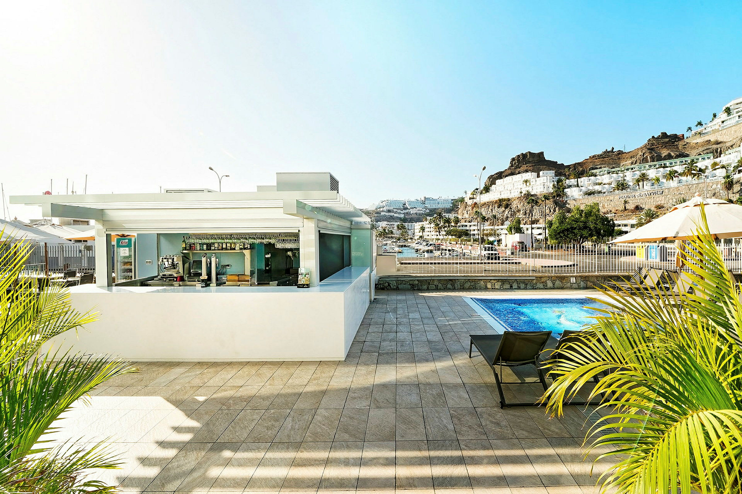 Bild på en bar vid en pool med utsikt över omgivande byggnader och berg. (AI-genererad text)