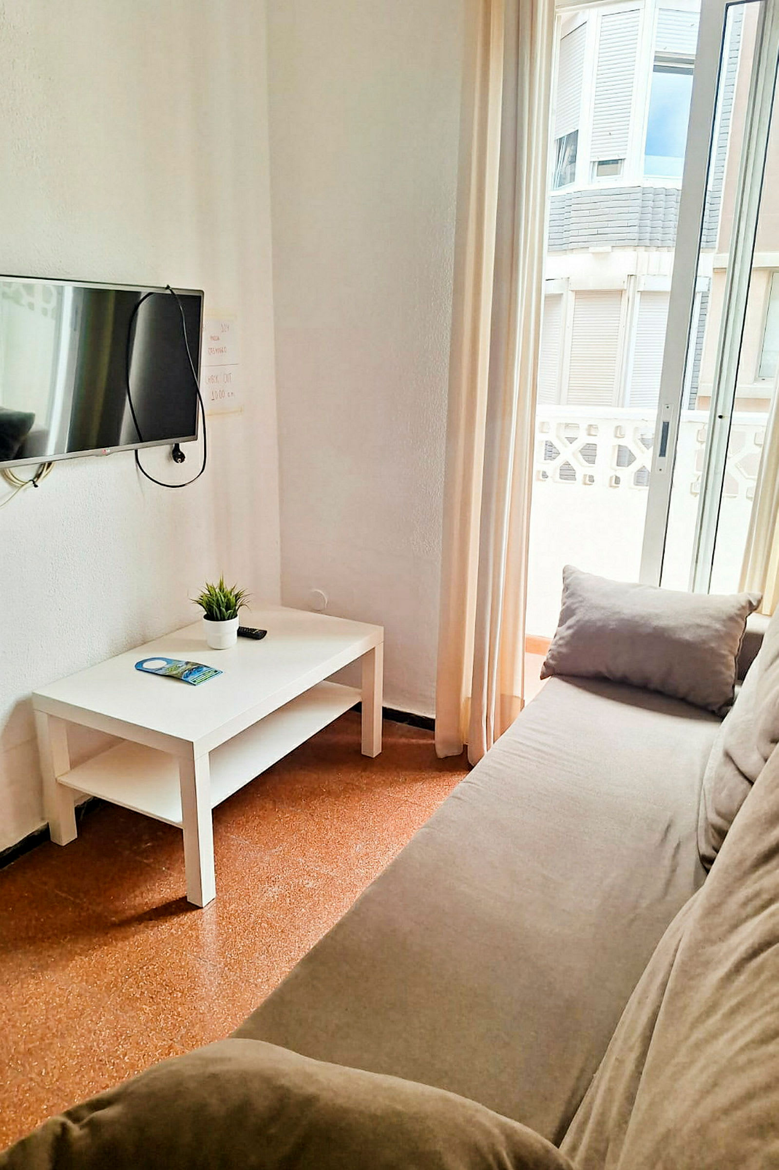 En soffa, ett soffbord med en växt och en broschyr, samt en väggmonterad TV i ett rum med fönster och ljusinsläpp. (AI-genererad text)