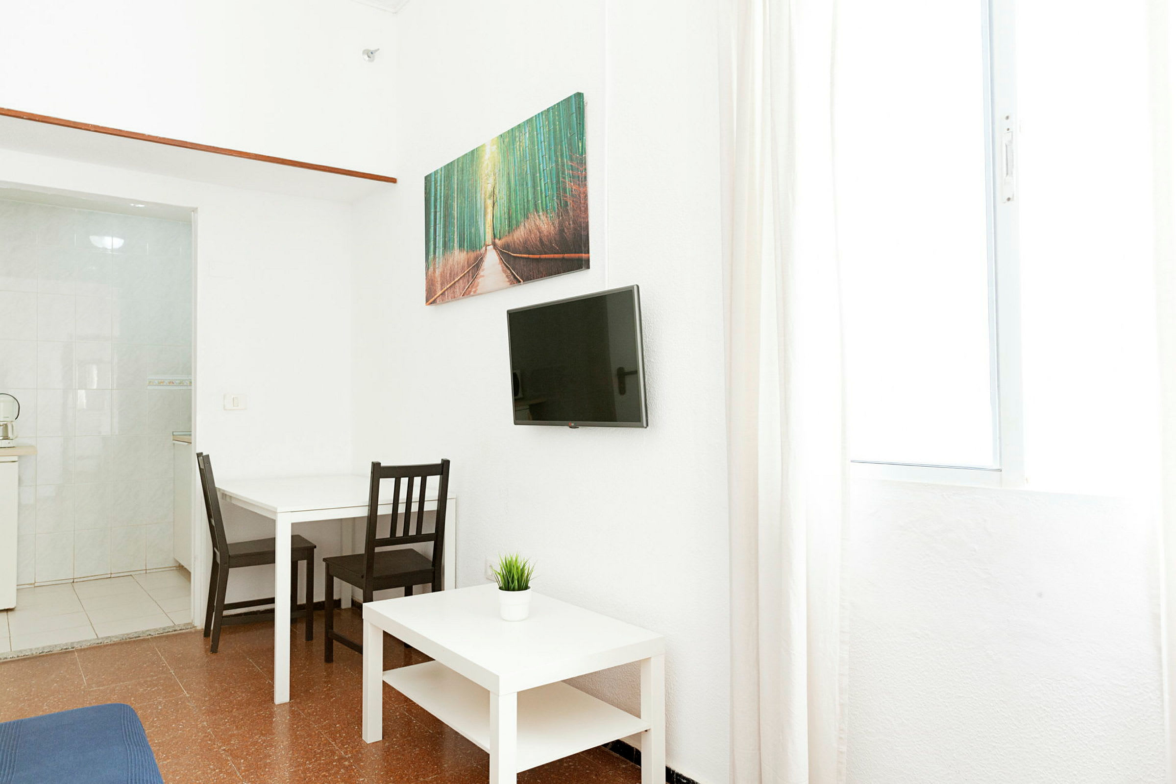 Ett litet rum med ett vitt bord och två stolar, en TV på väggen och ett litet bord med en krukväxt. (AI-genererad text)