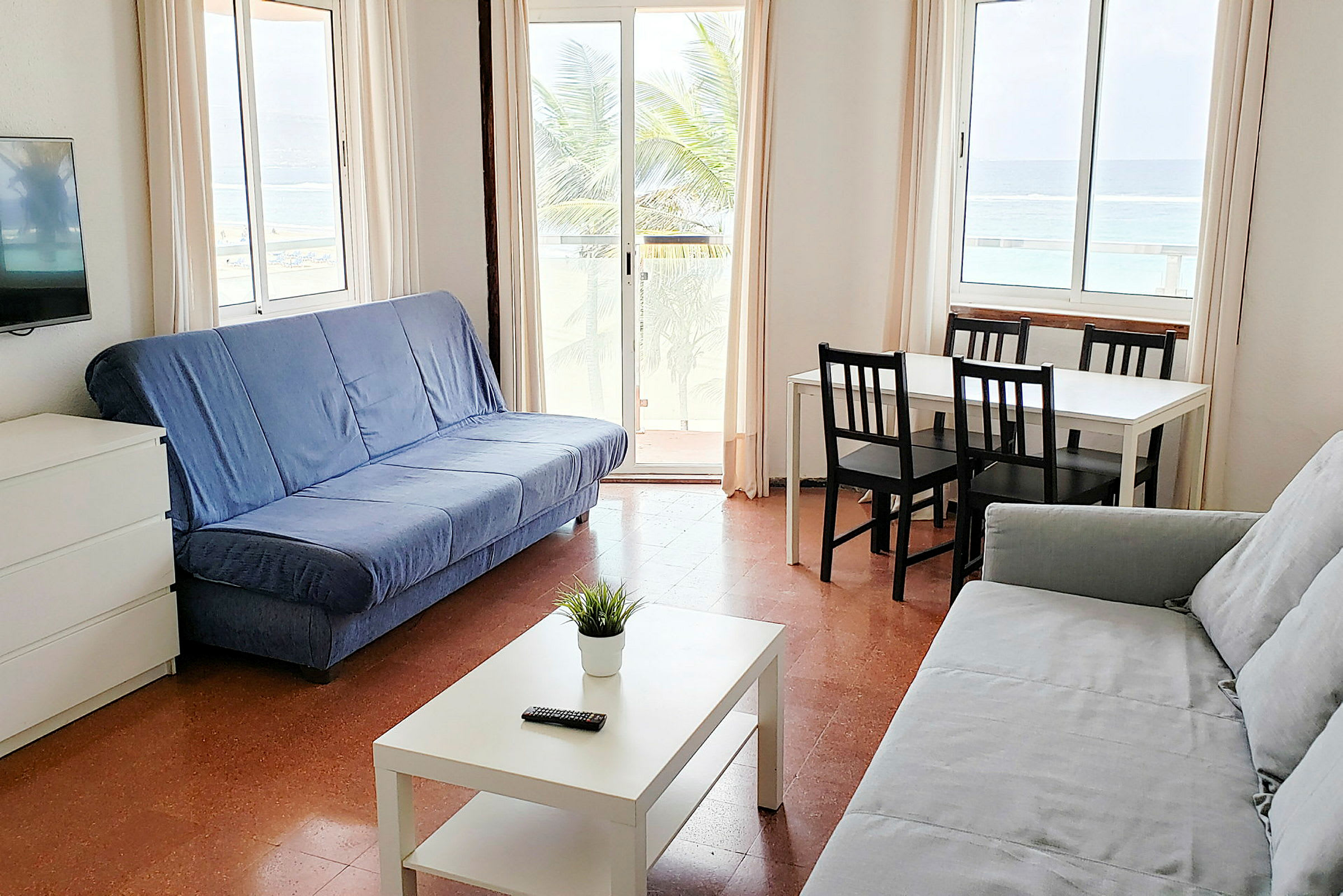 Ett vardagsrum med två soffor, ett soffbord, en TV och ett matbord med fyra stolar. Utsikt mot stranden kan ses genom fönstren. (AI-genererad text)