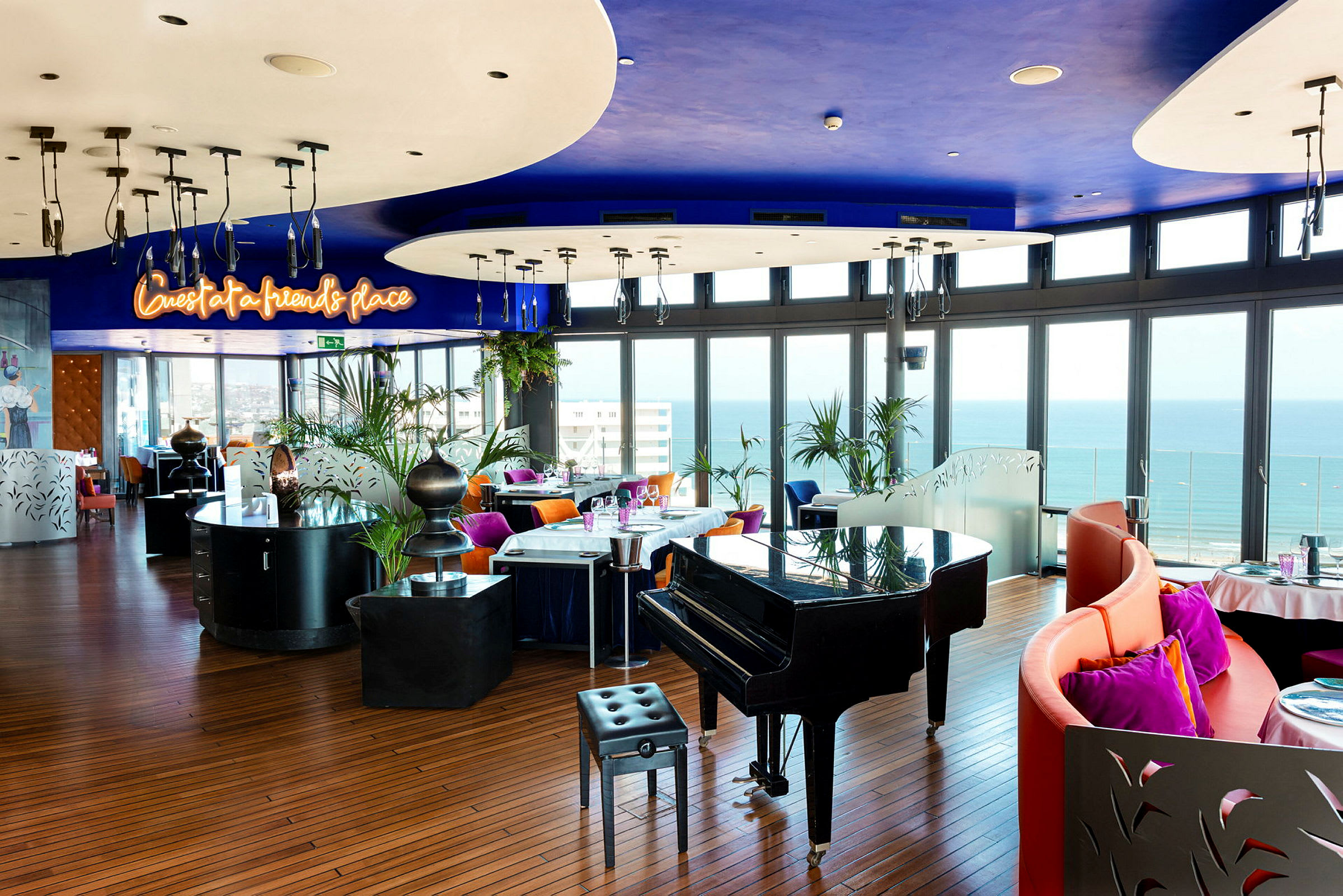 En restaurang med modern inredning, färgglada stolar och ett piano med utsikt över havet från stora fönster. (AI-genererad text)