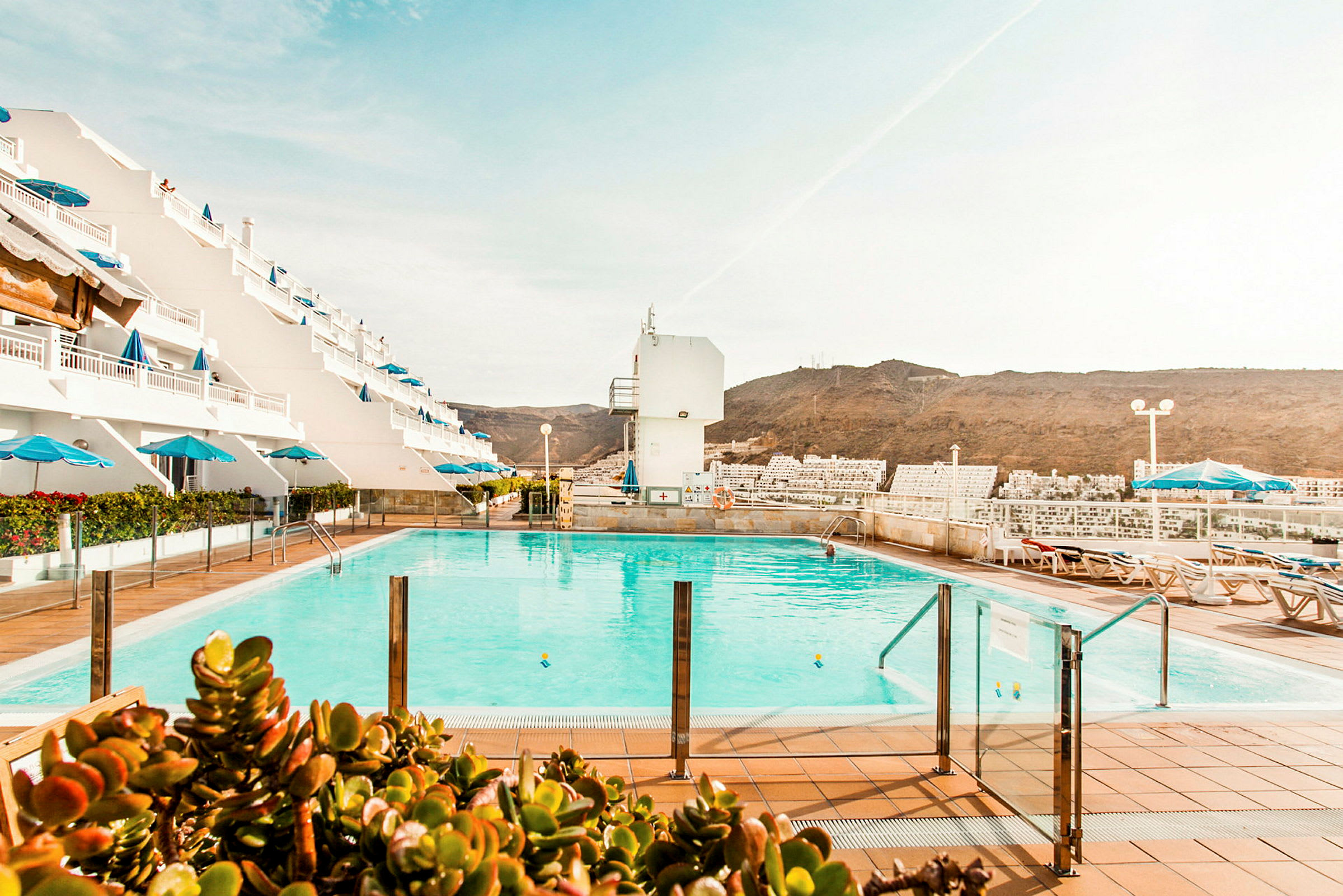 Poolområde med solstolar och parasoll, omgiven av ett hotell med vita fasader och utsikt över bergen. (AI-genererad text)