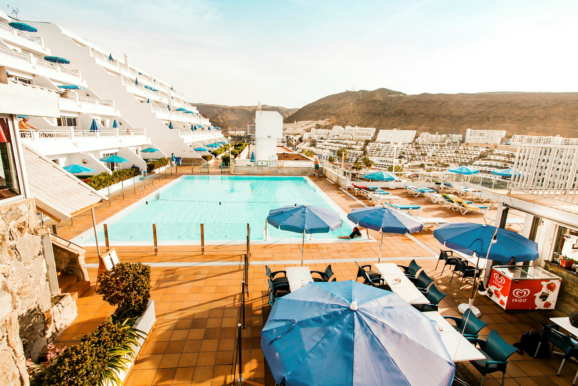 Bild av en anläggning med pool, solstolar och blå parasoller framför ett hotell med terrasser. (AI-genererad text)