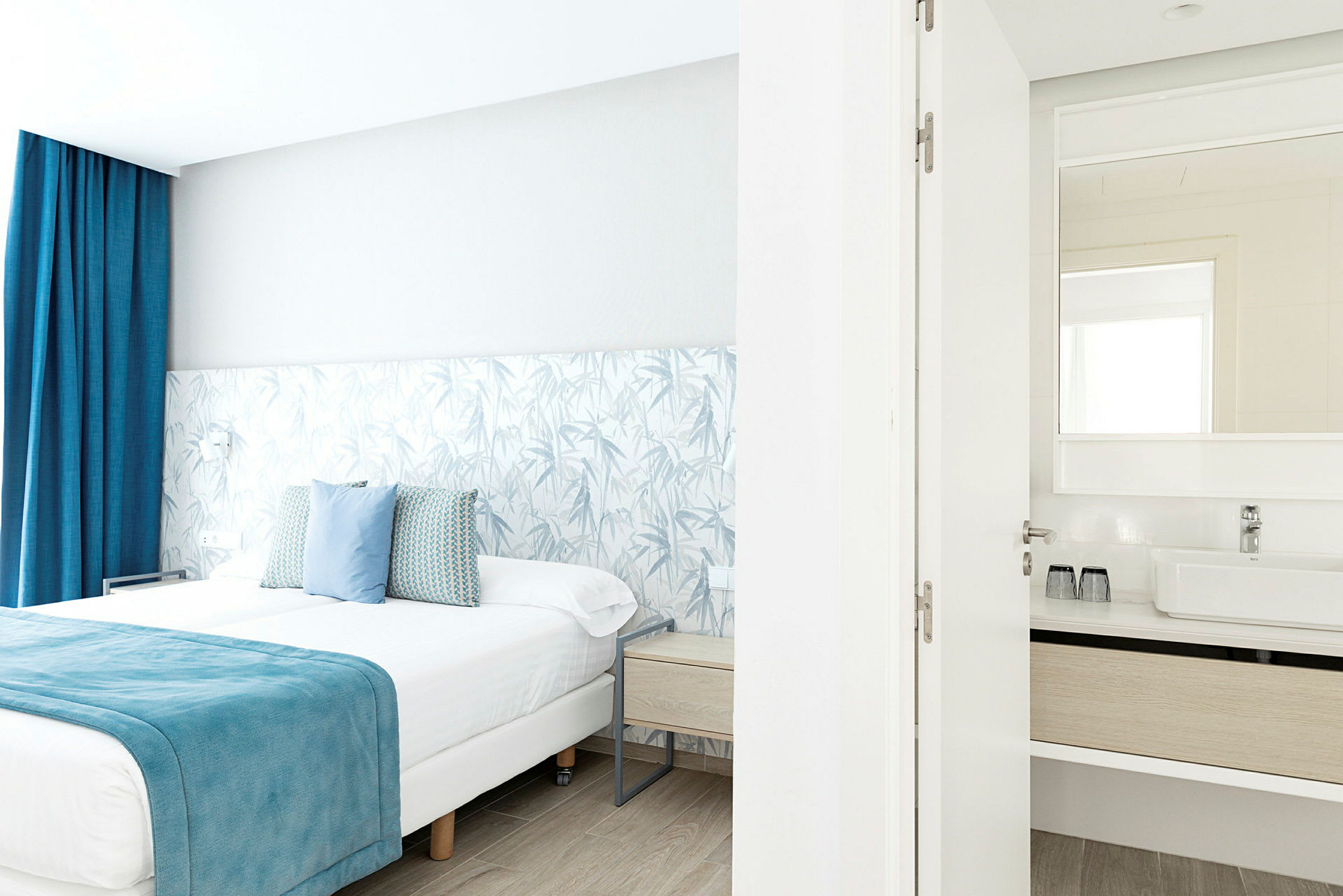 Ett ljust hotellrum med en dubbelsäng med blå detaljer och en anslutande badrum med synlig spegel. (AI-genererad text)
