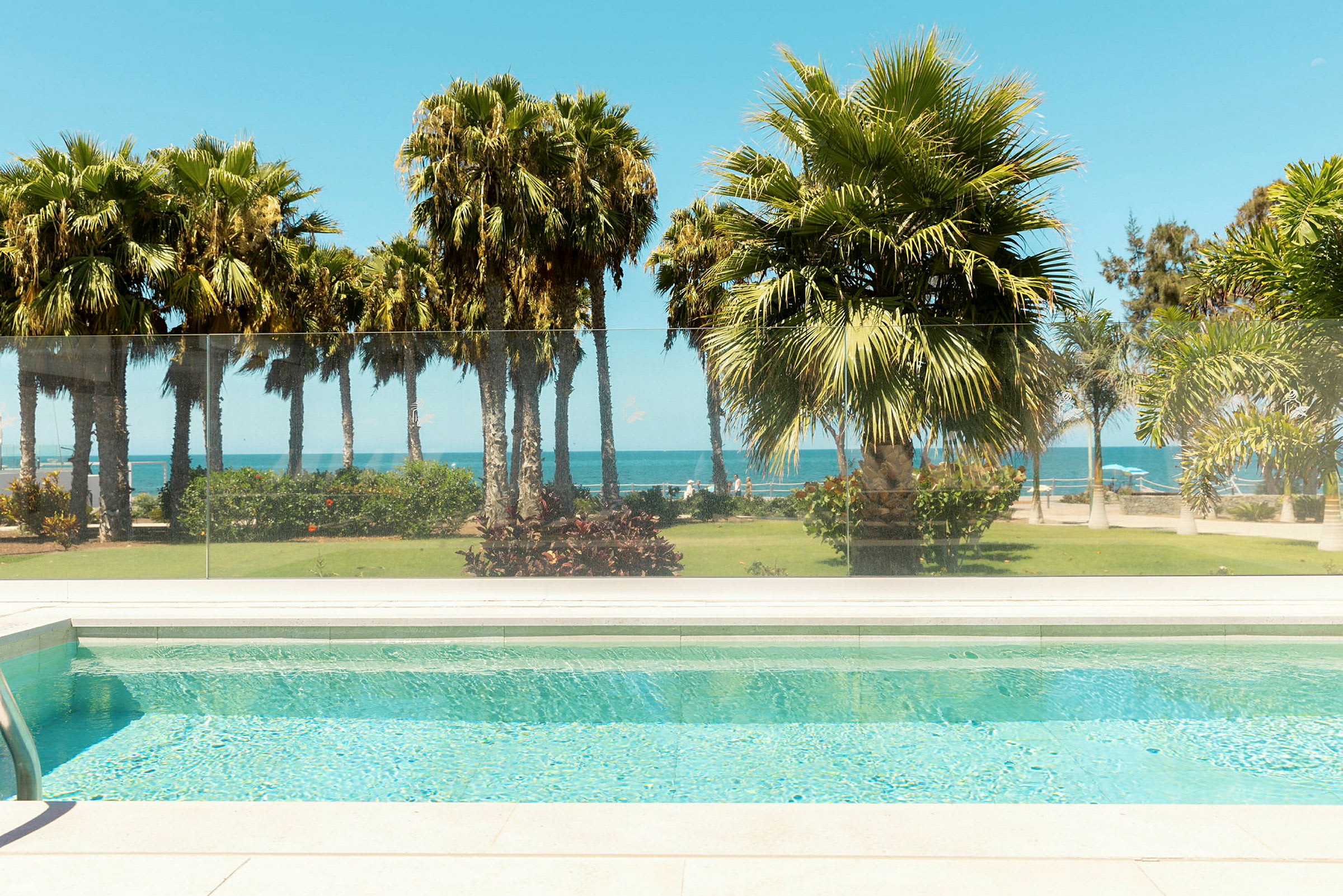 Utsikt från en pool mot en trädgård med palmer och havet i bakgrunden. (AI-genererad text)