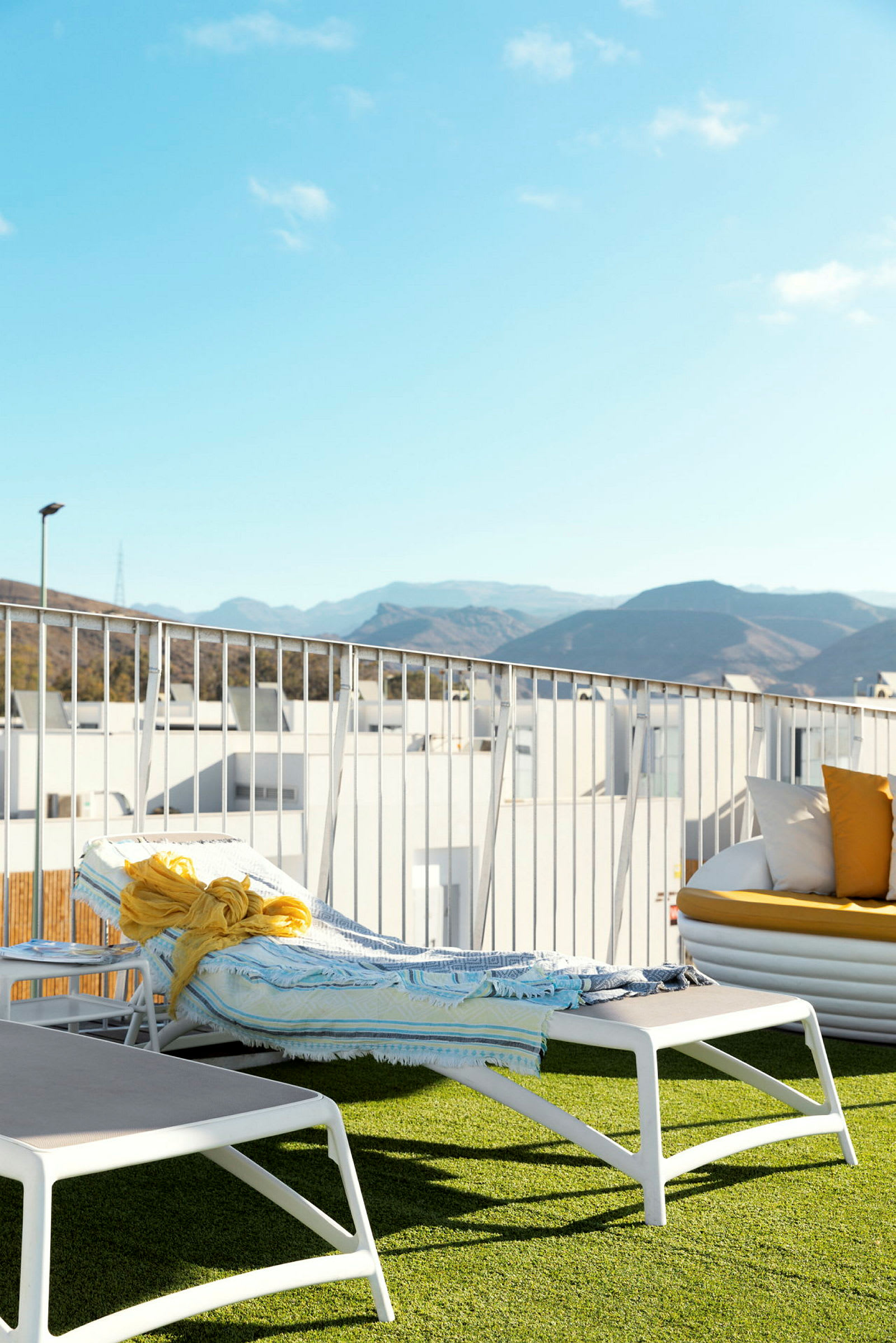 En solstol med en handduk och en gul sjal ligger på en terrass med konstgräs, med berg och blå himmel i bakgrunden. (AI-genererad text)