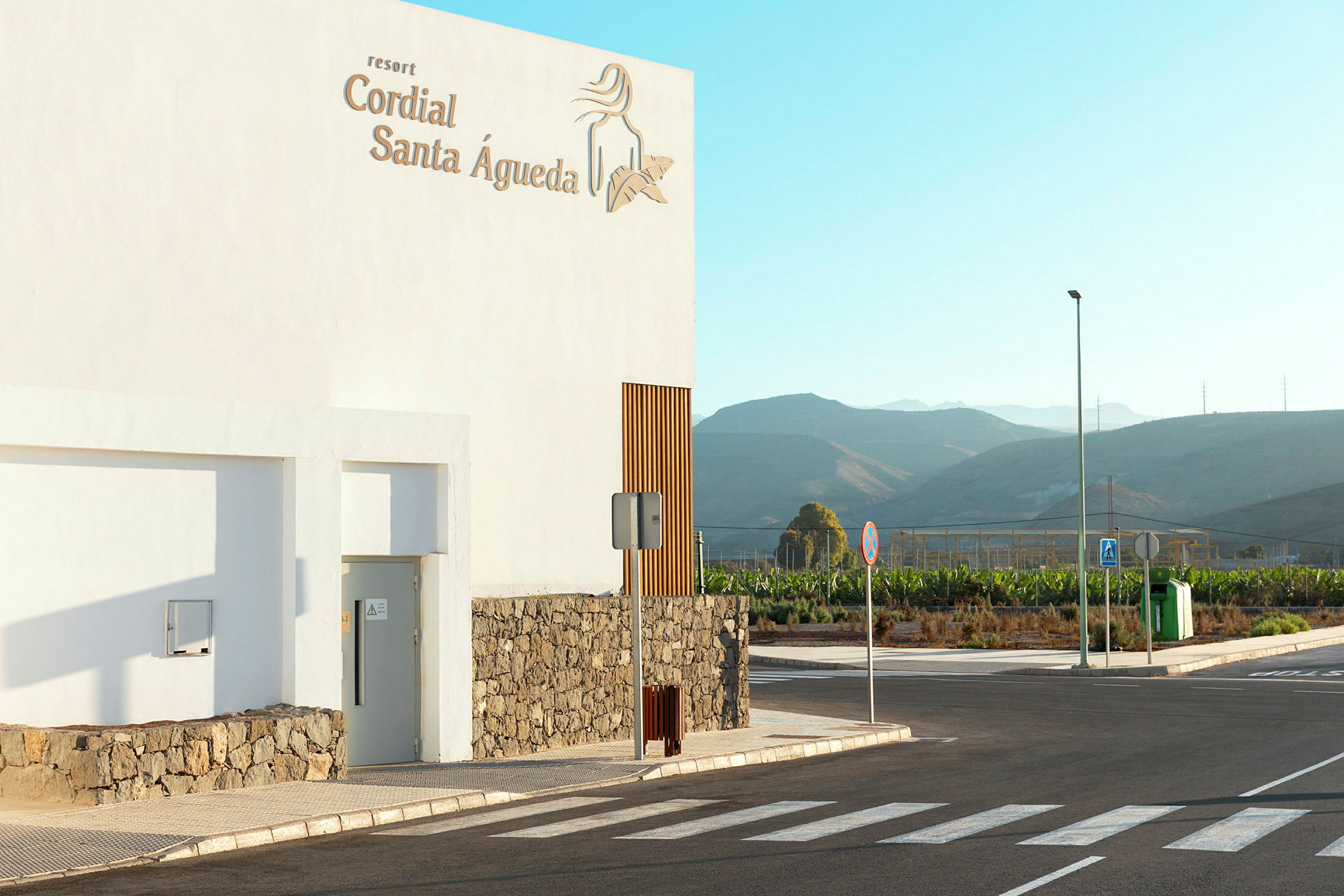 Vit byggnad med skylten 'Cordial Santa Águeda', framför ett landskap med berg i bakgrunden. (AI-genererad text)