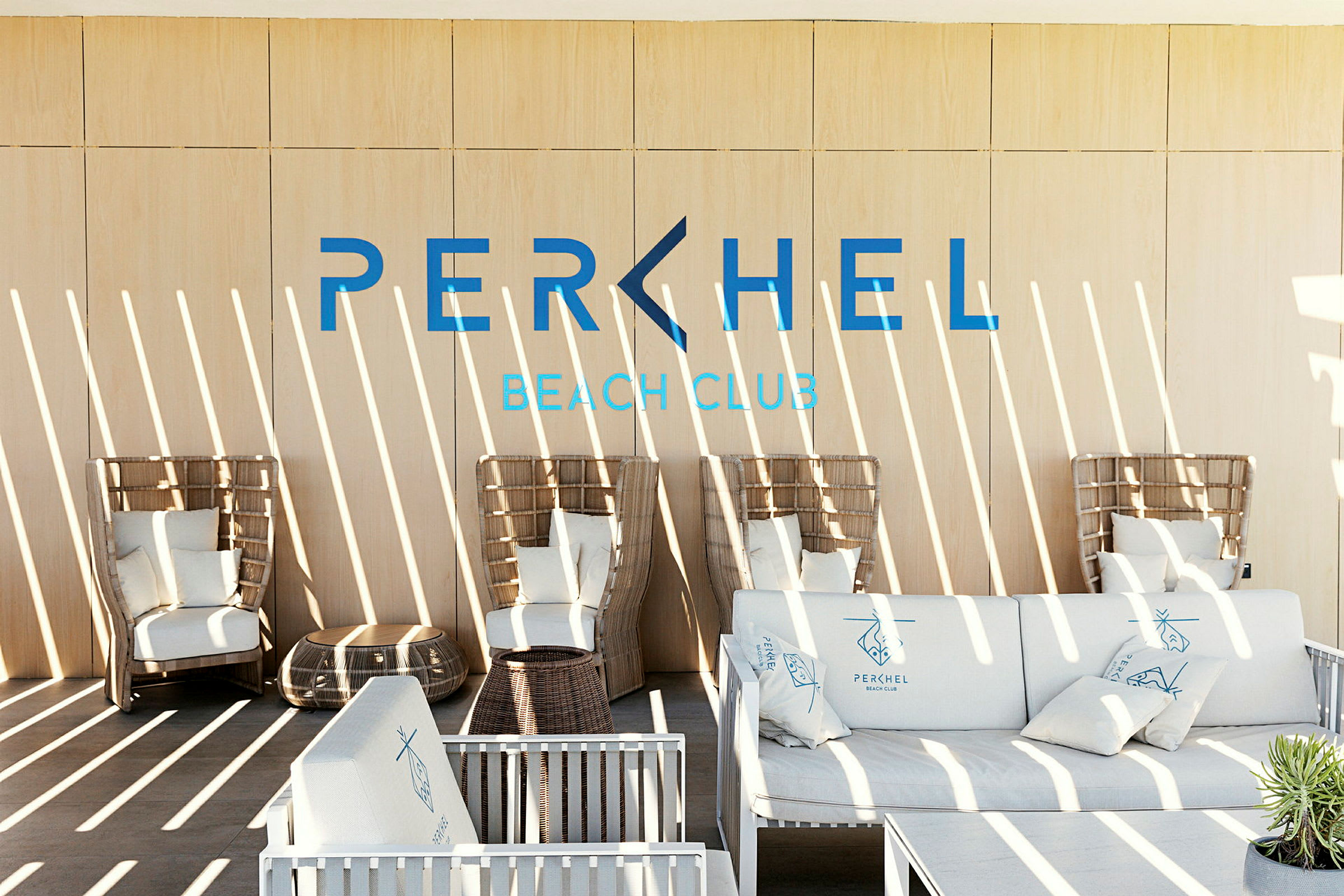 Ett modernt loungeområde med korgfåtöljer och soffor med kuddar samt texten 'Perchel Beach Club' på väggen. (AI-genererad text)
