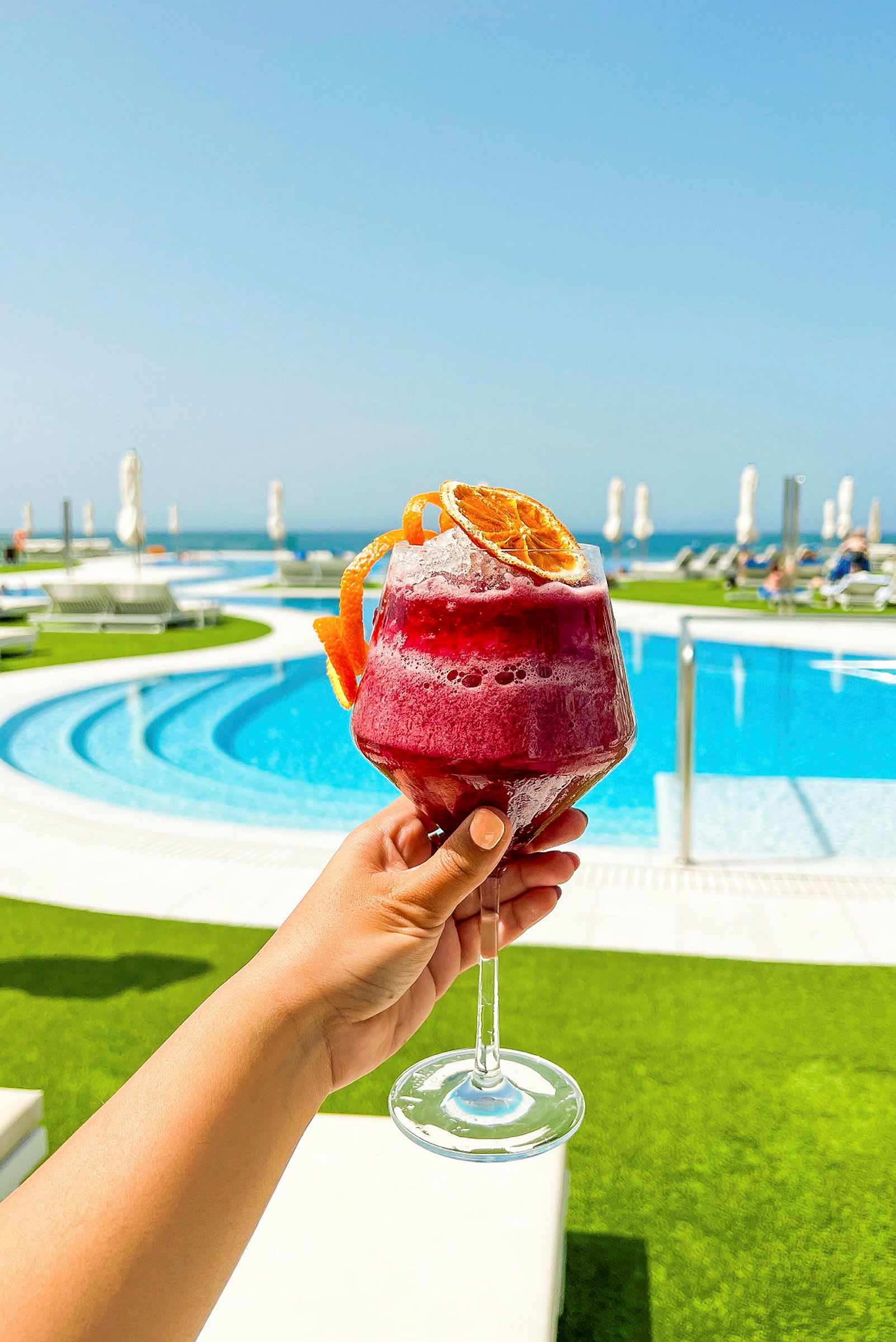 Ett glas med röd dryck dekorerat med torkade apelsinskivor hålls upp framför en swimmingpool utomhus med solsängar och ett blått hav i bakgrunden. (AI-genererad text)