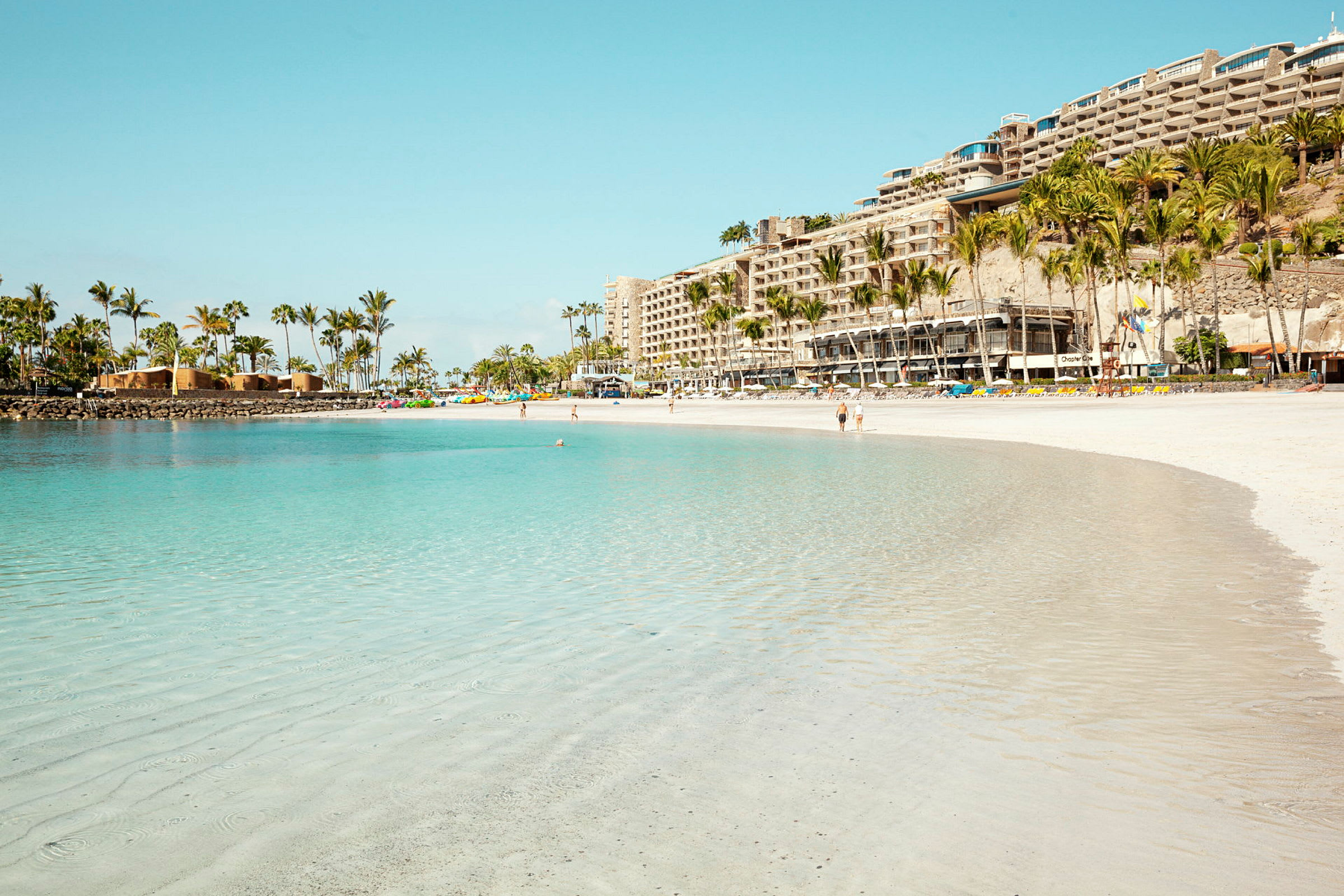 En sandstrand med turkost vatten och hotell samt palmer i bakgrunden. (AI-genererad text)