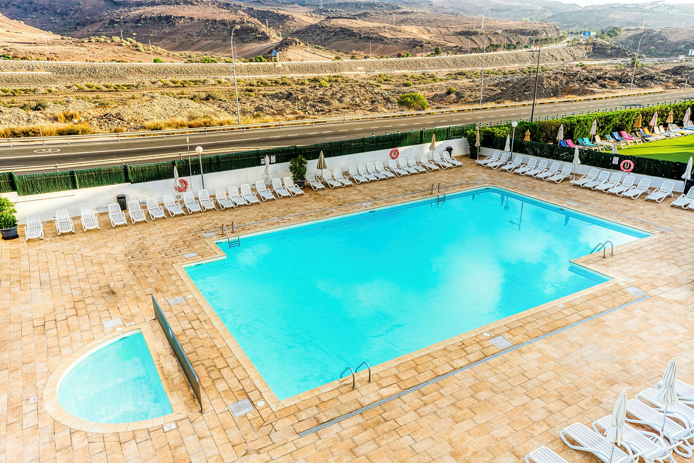 En swimmingpool med omgivande solstolar och höjd landskap i bakgrunden. (AI-genererad text)