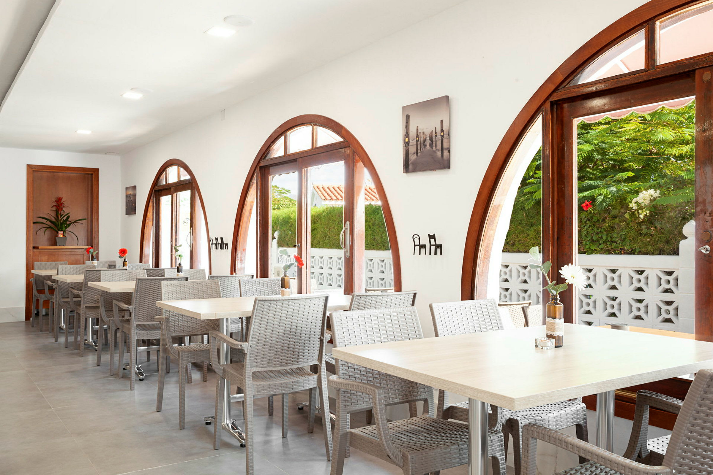 Restaurang med fönster med bågform och bord med stolar. Växter utanför synliga genom fönstren. (AI-genererad text)