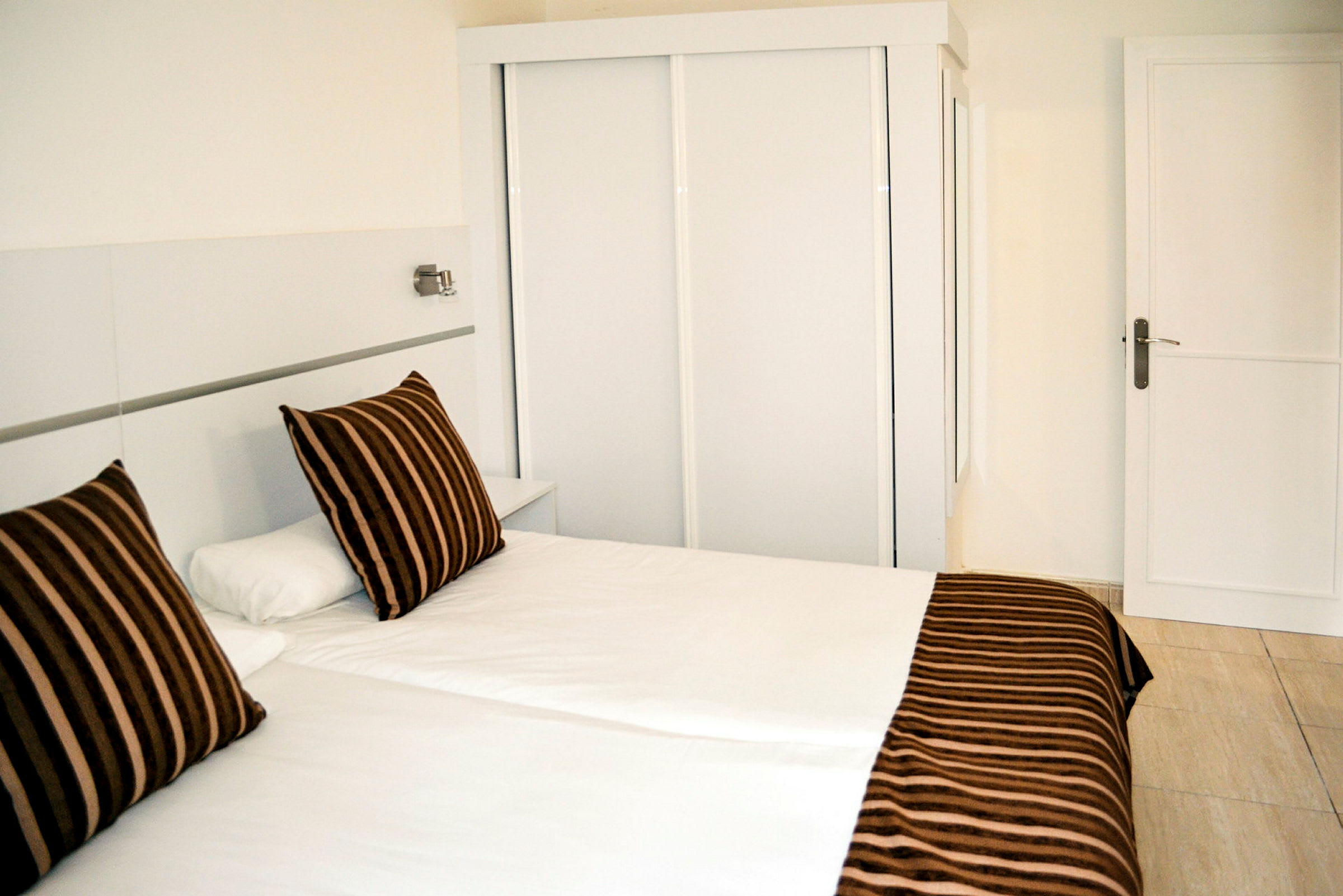 Ett hotellrum med en vit säng och kuddar med brunt randigt mönster framför en vit garderob. (AI-genererad text)