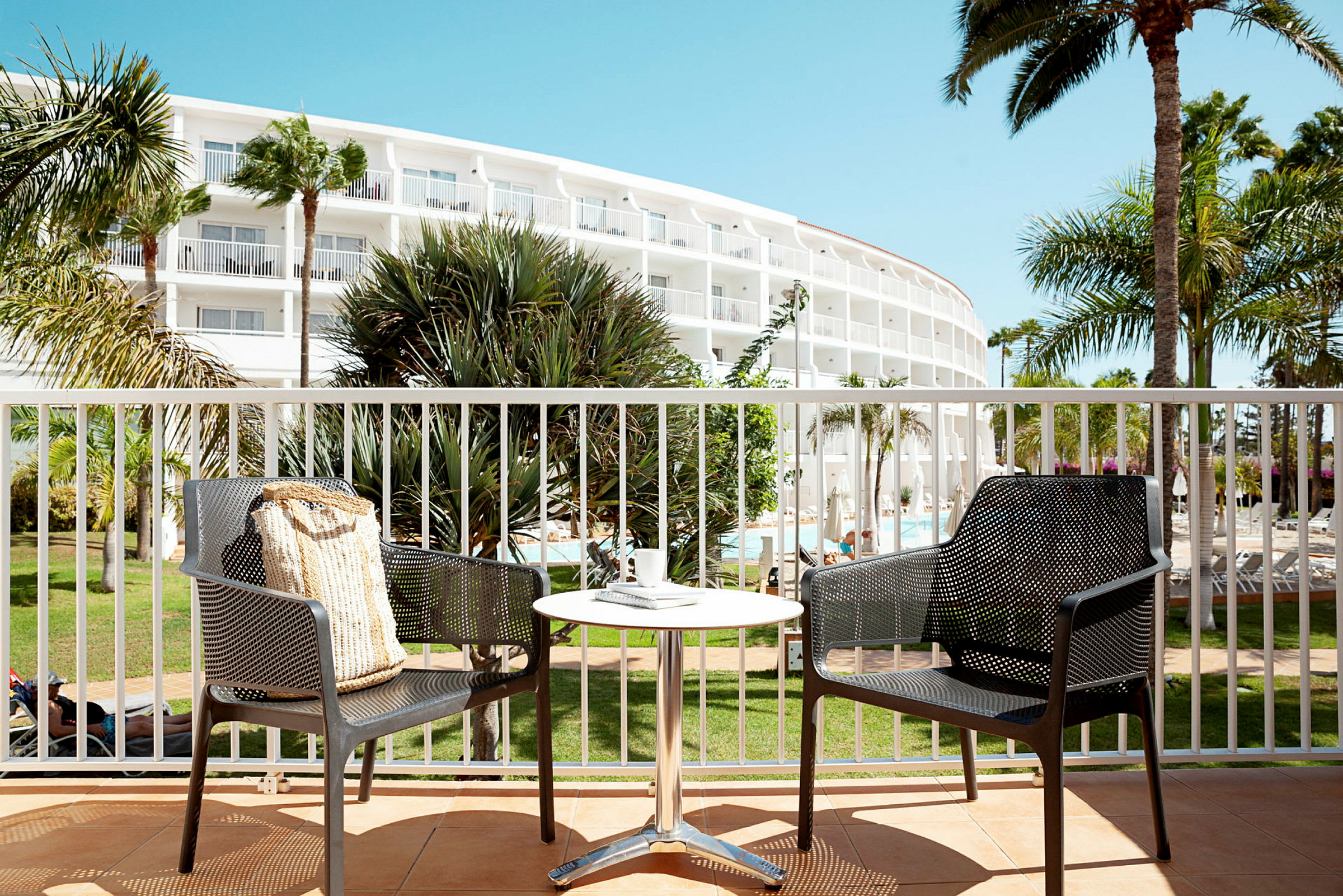 Två stolar och ett bord på en terrass med utsikt över en hotellbyggnad och omgivande palmer. (AI-genererad text)