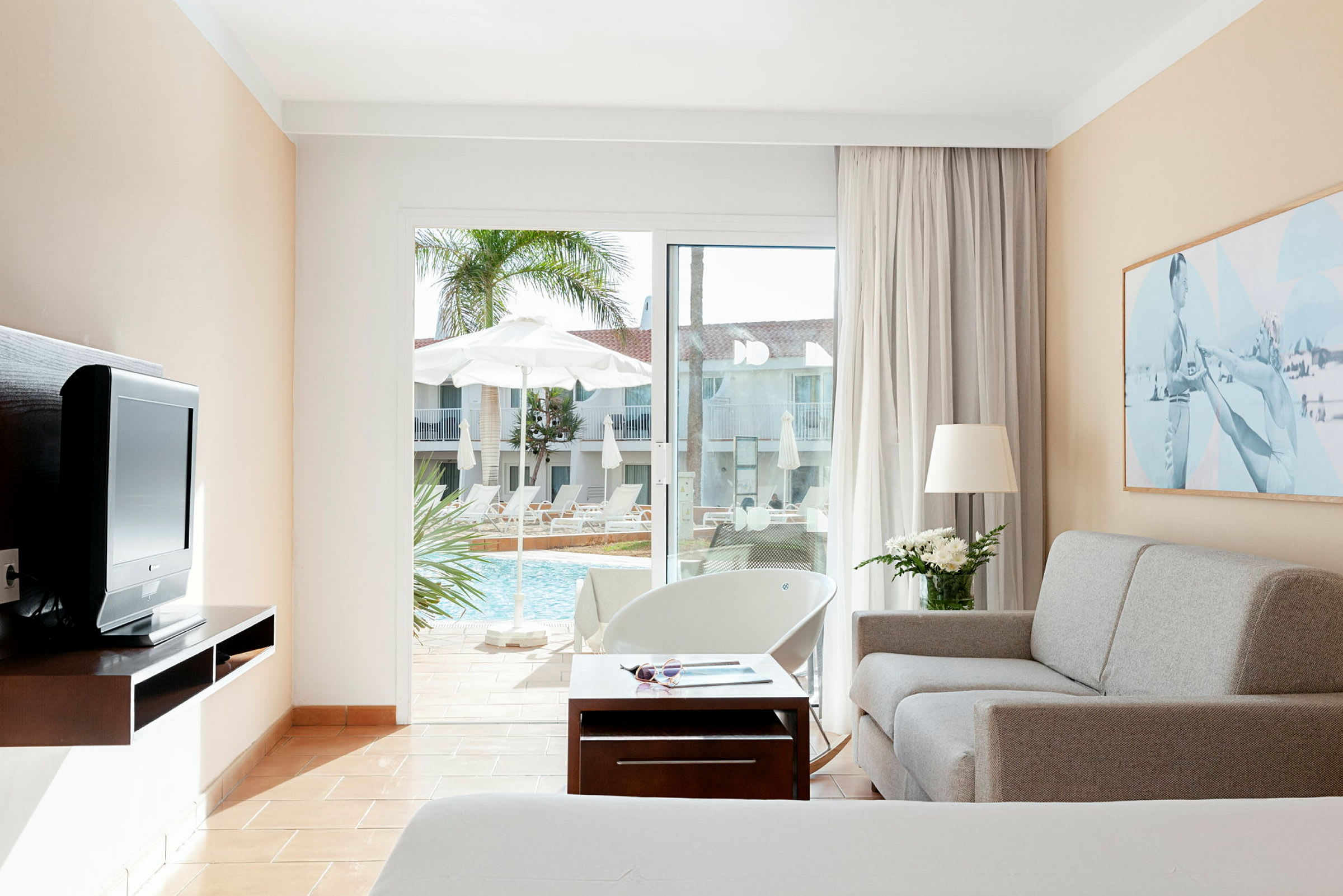 En hotellrumsinteriör med soffa, bord och TV, samt utsikt till poolområdet genom en glasad altandörr. (AI-genererad text)