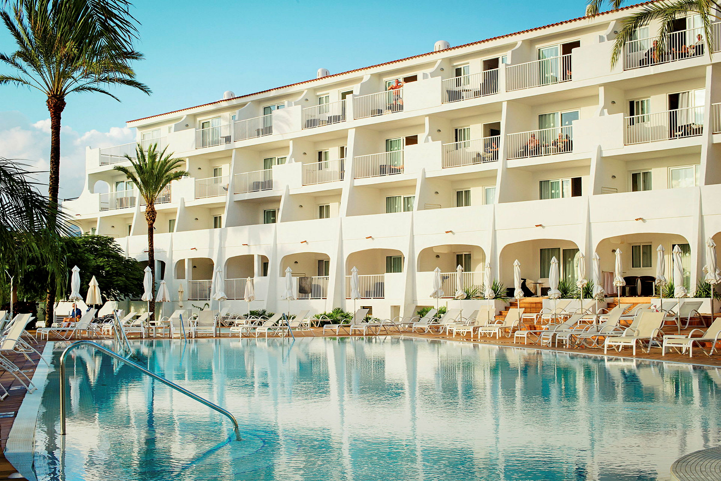 Hotell med vit fasad och utomhuspool omgiven av solstolar. (AI-genererad text)