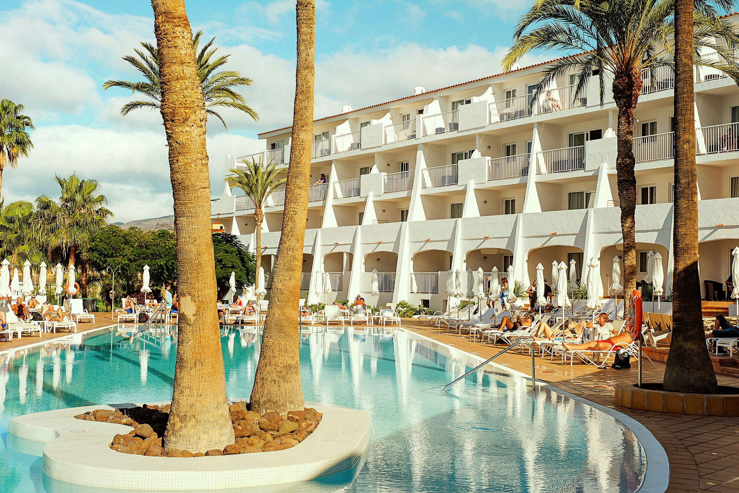 Hotellbyggnad vid en pool, omgiven av solstolar och palmer. (AI-genererad text)
