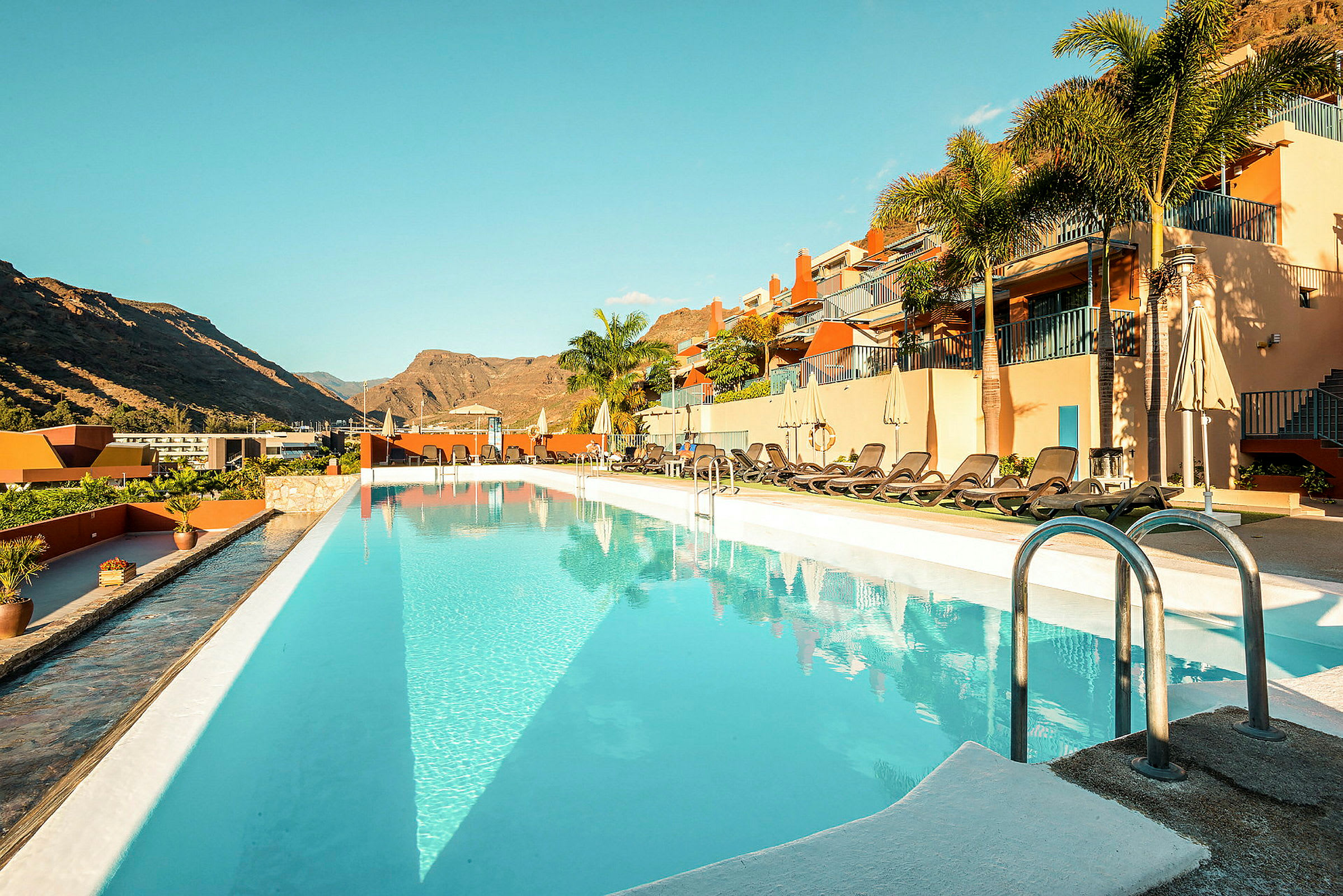 Utomhuspool vid ett hotell med solstolar och berg i bakgrunden. (AI-genererad text)